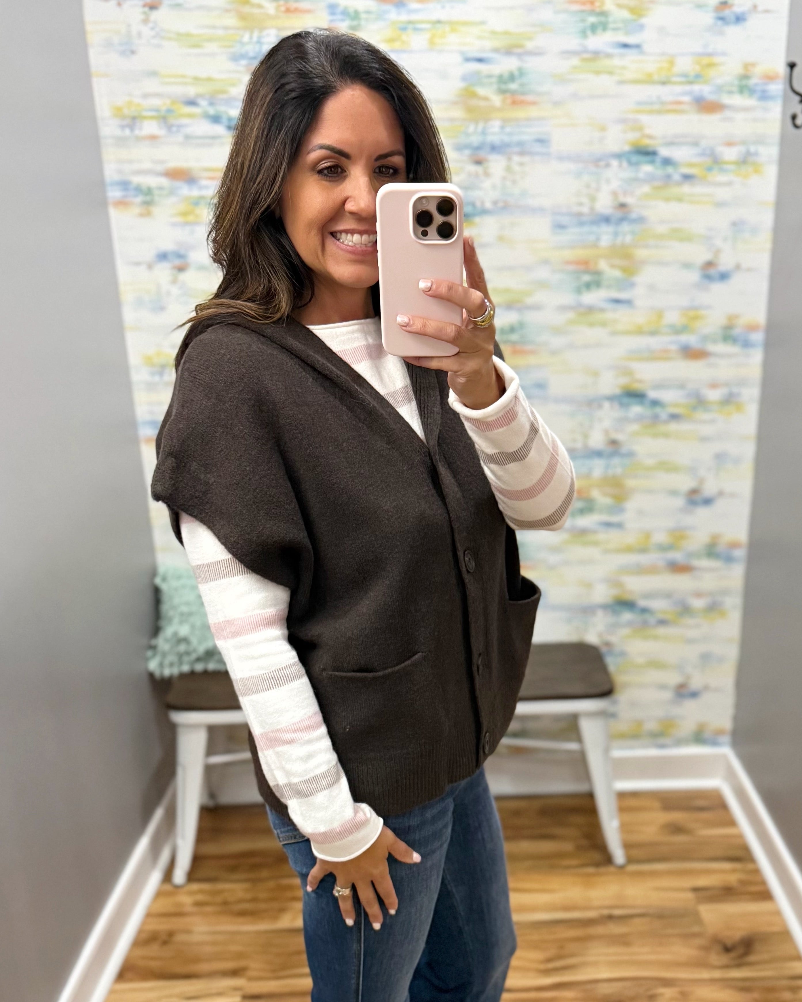 A woman takes a mirror selfie in a fitting room wearing a dark brown sleeveless button-front cardigan over a white and pink striped long-sleeve shirt and blue jeans.