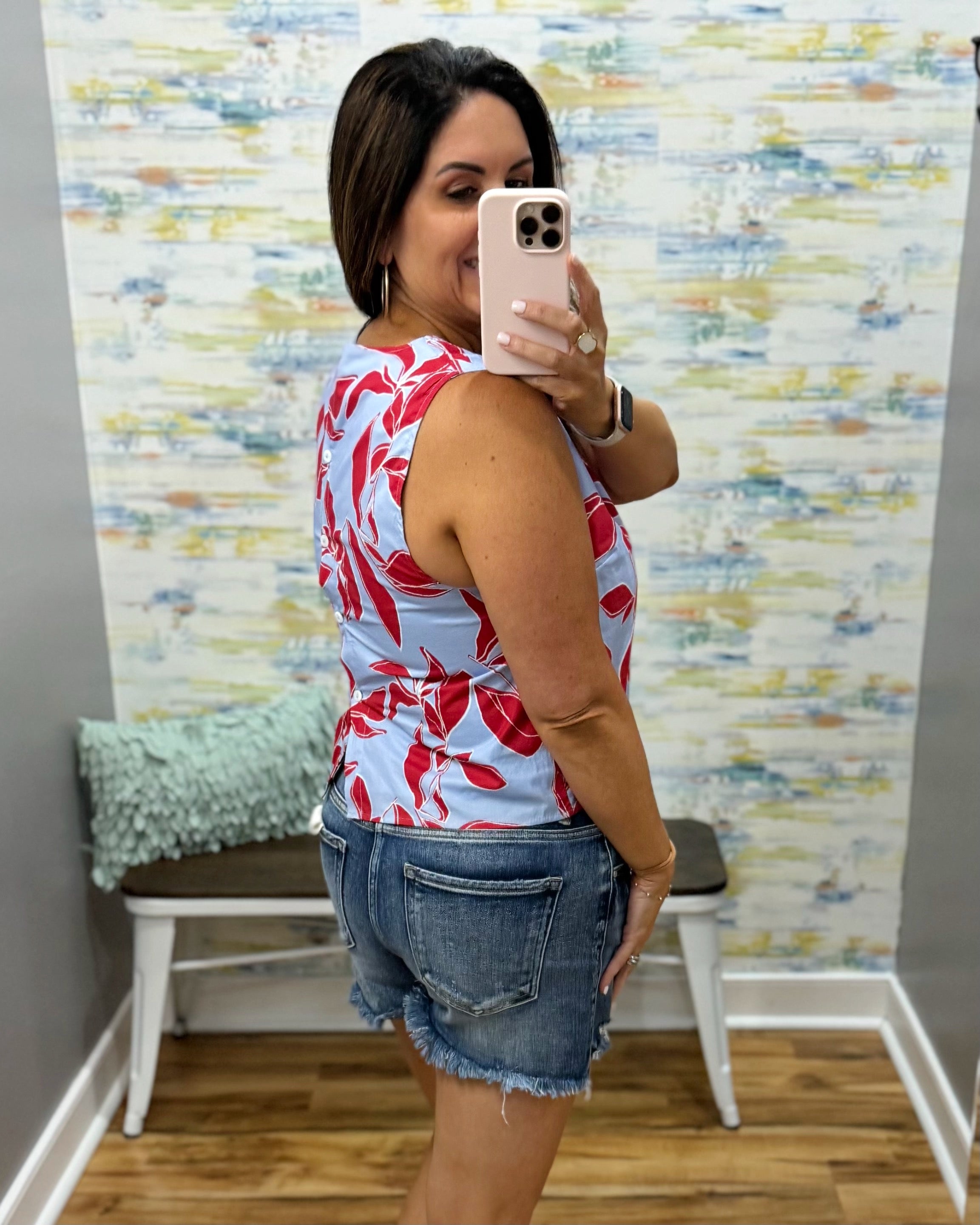 Woman taking a mirror selfie wearing a floral top and denim shorts in a room with colorful wall art.