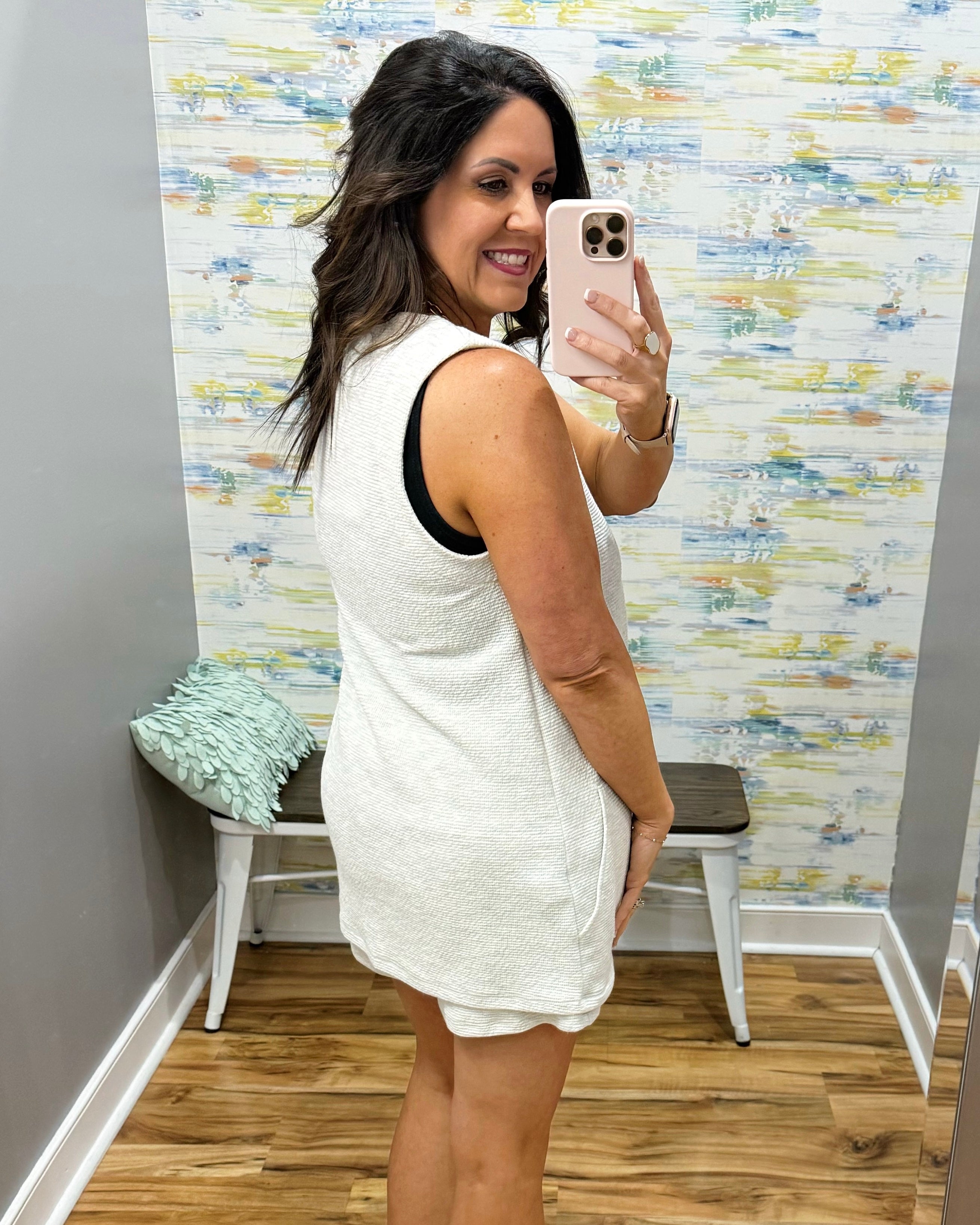 Woman taking a mirror selfie wearing a white sleeveless dress in a room with colorful wall paper.