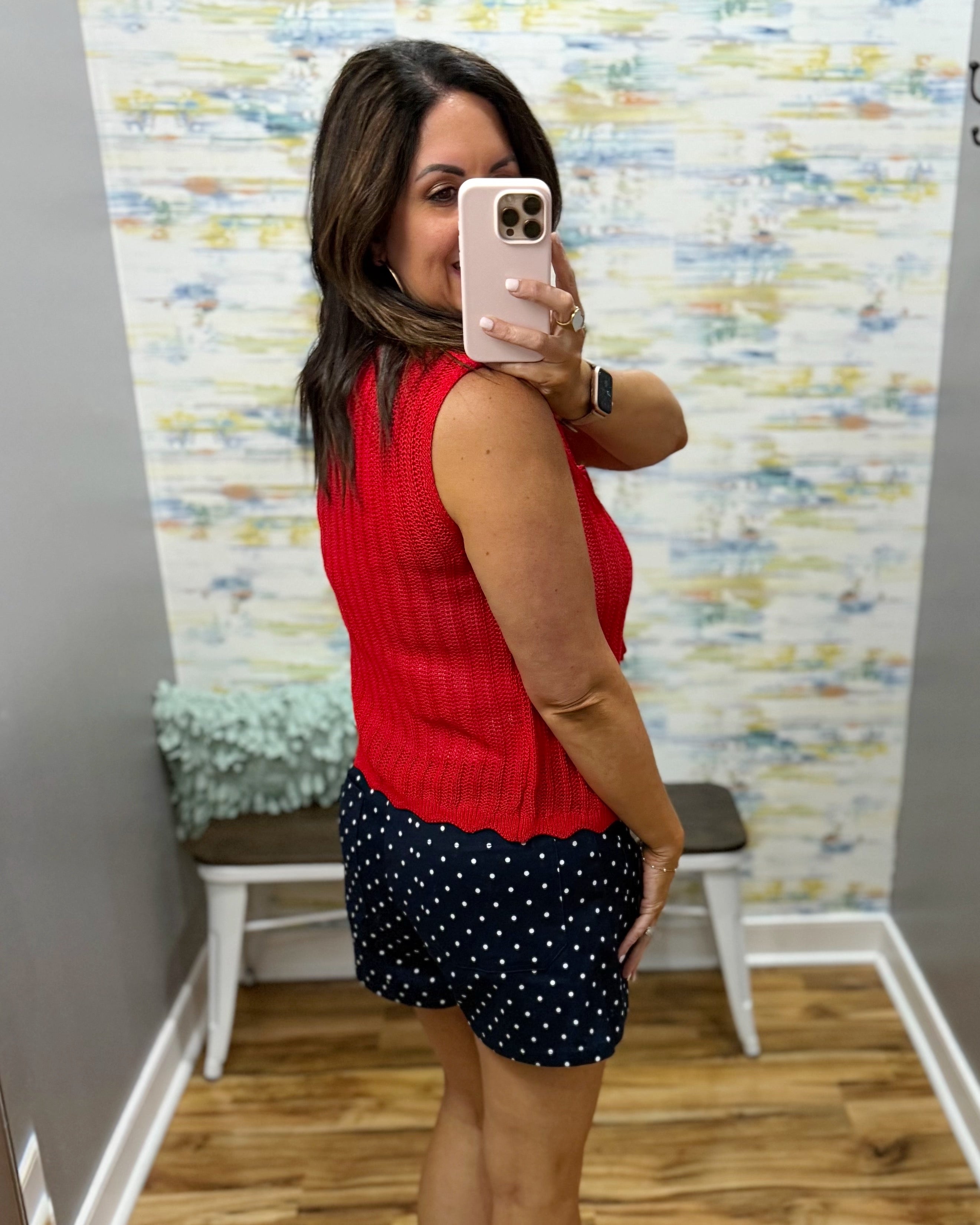 Woman taking a mirror selfie wearing a red sleeveless top and black polka dot shorts in a room with a textured wall.