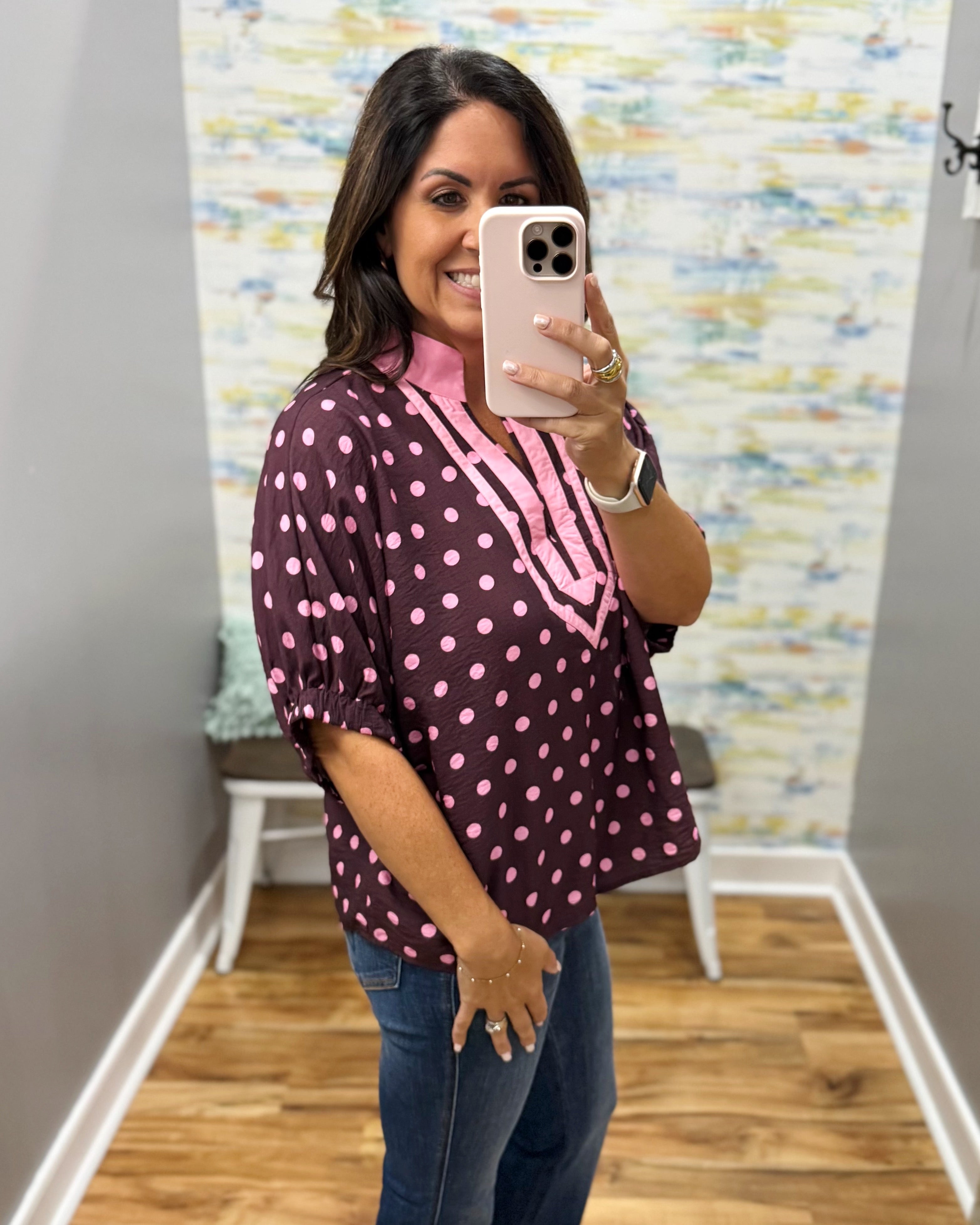 A woman takes a mirror selfie wearing a dark plum purple blouse with pink polka dots and a pink V-neckline trim, paired with blue jeans.