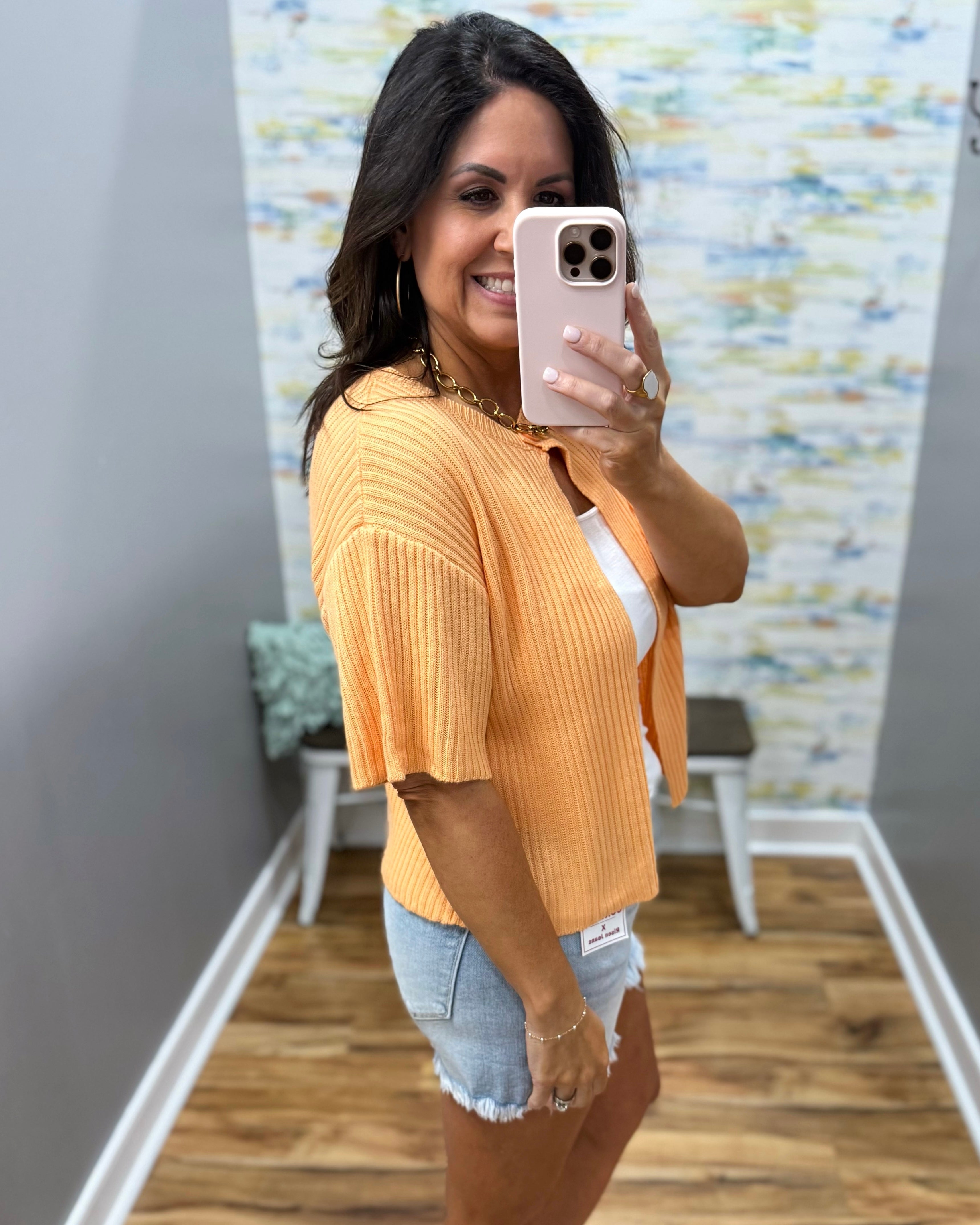 Woman taking a mirror selfie wearing a yellow cardigan and denim shorts in a room with a textured wall.