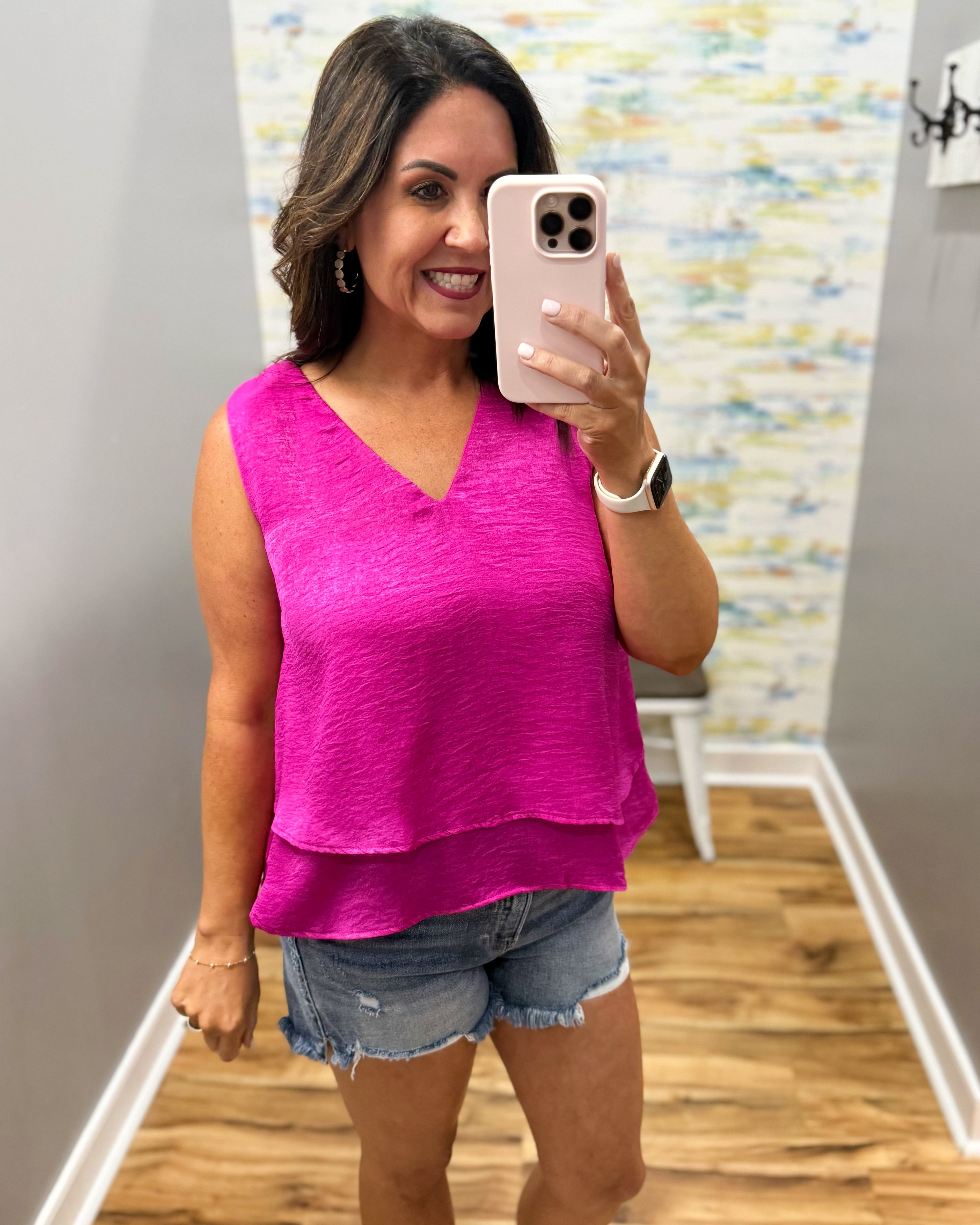 Woman taking a mirror selfie wearing a bright pink top and denim shorts in a room with a patterned wall.