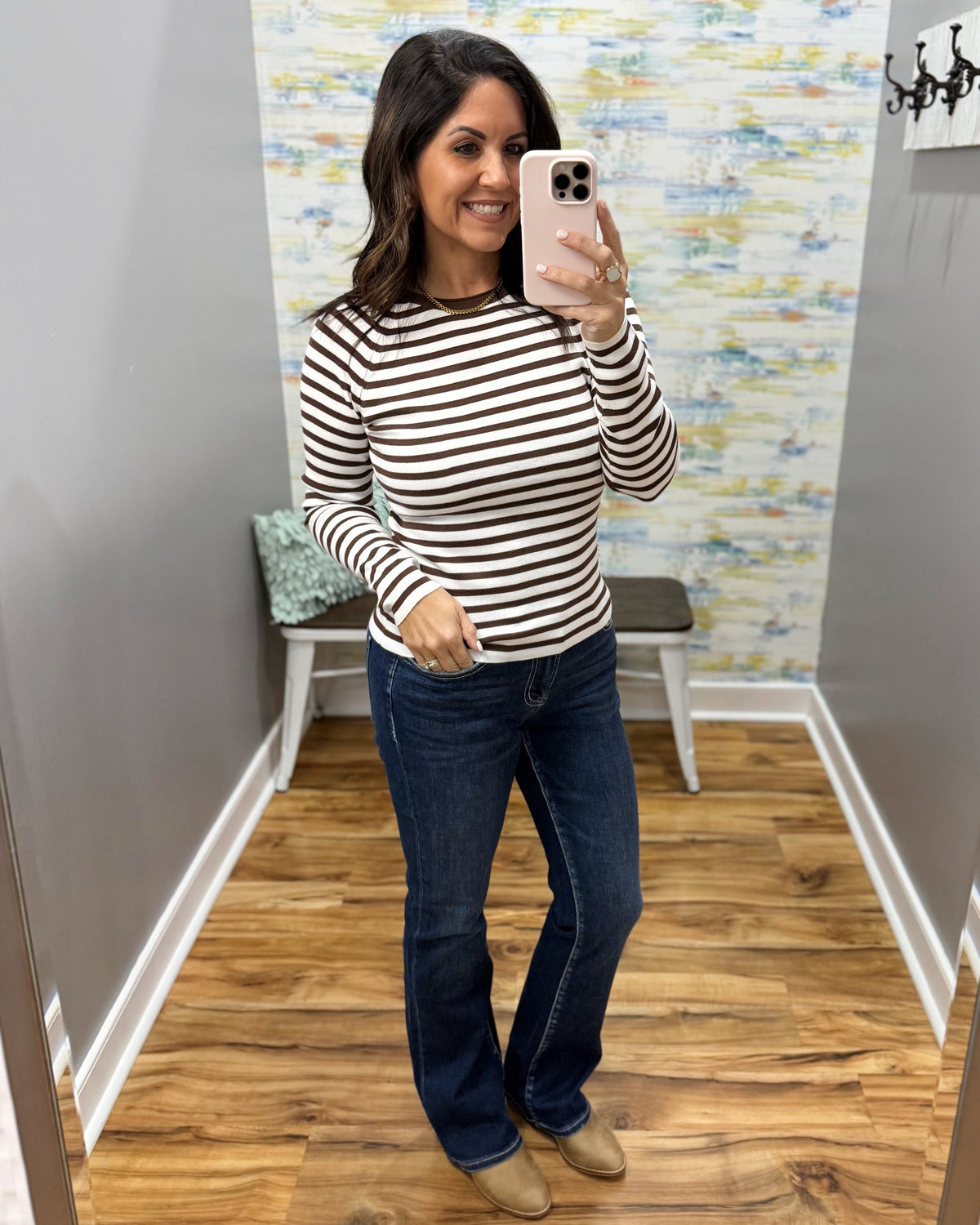 A woman takes a mirror selfie in a fitting room wearing a fitted long-sleeve top with horizontal black and off-white stripes, paired with dark blue jeans and tan shoes.