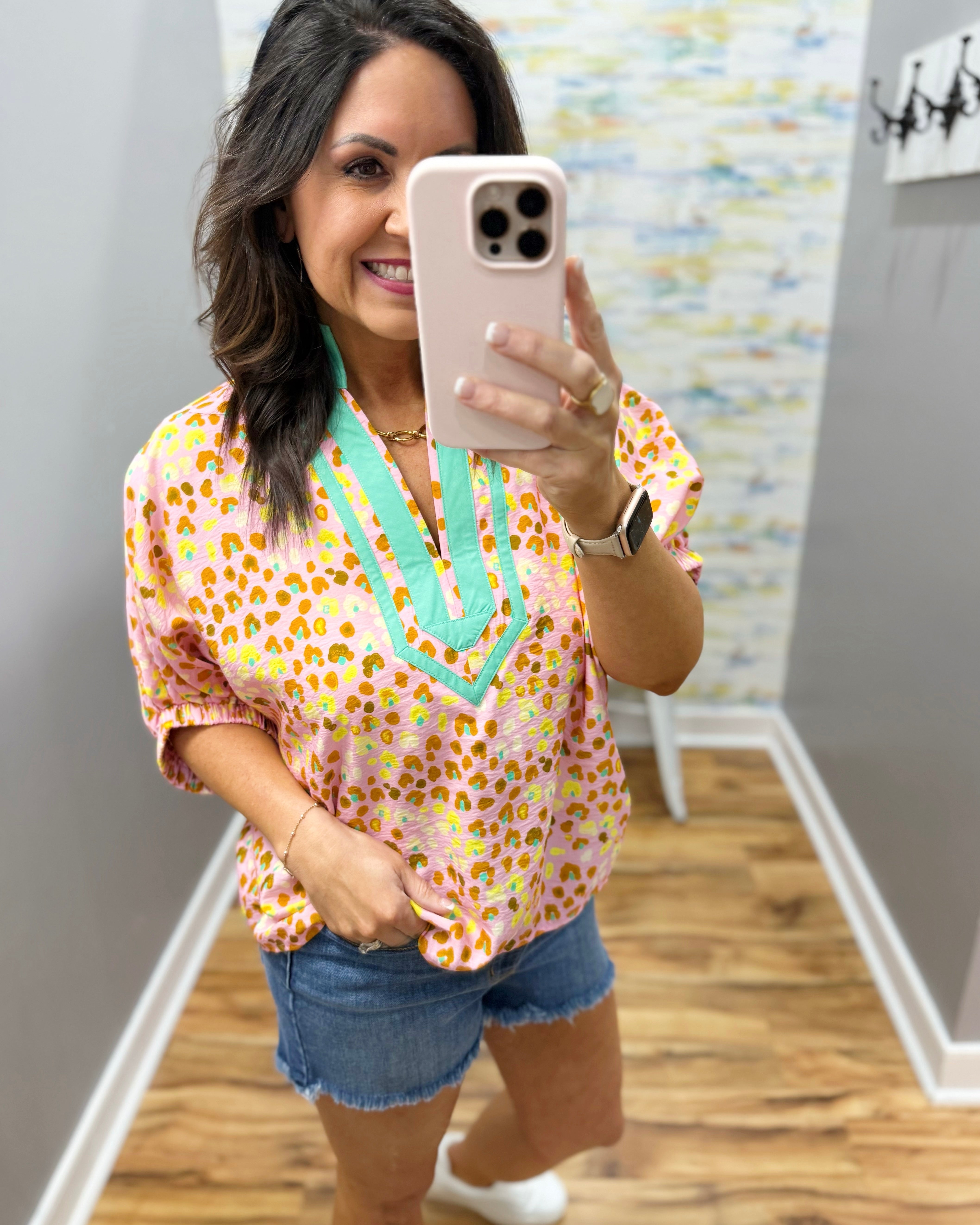 Woman taking a mirror selfie wearing a colorful blouse and denim shorts in a room with a patterned wall.