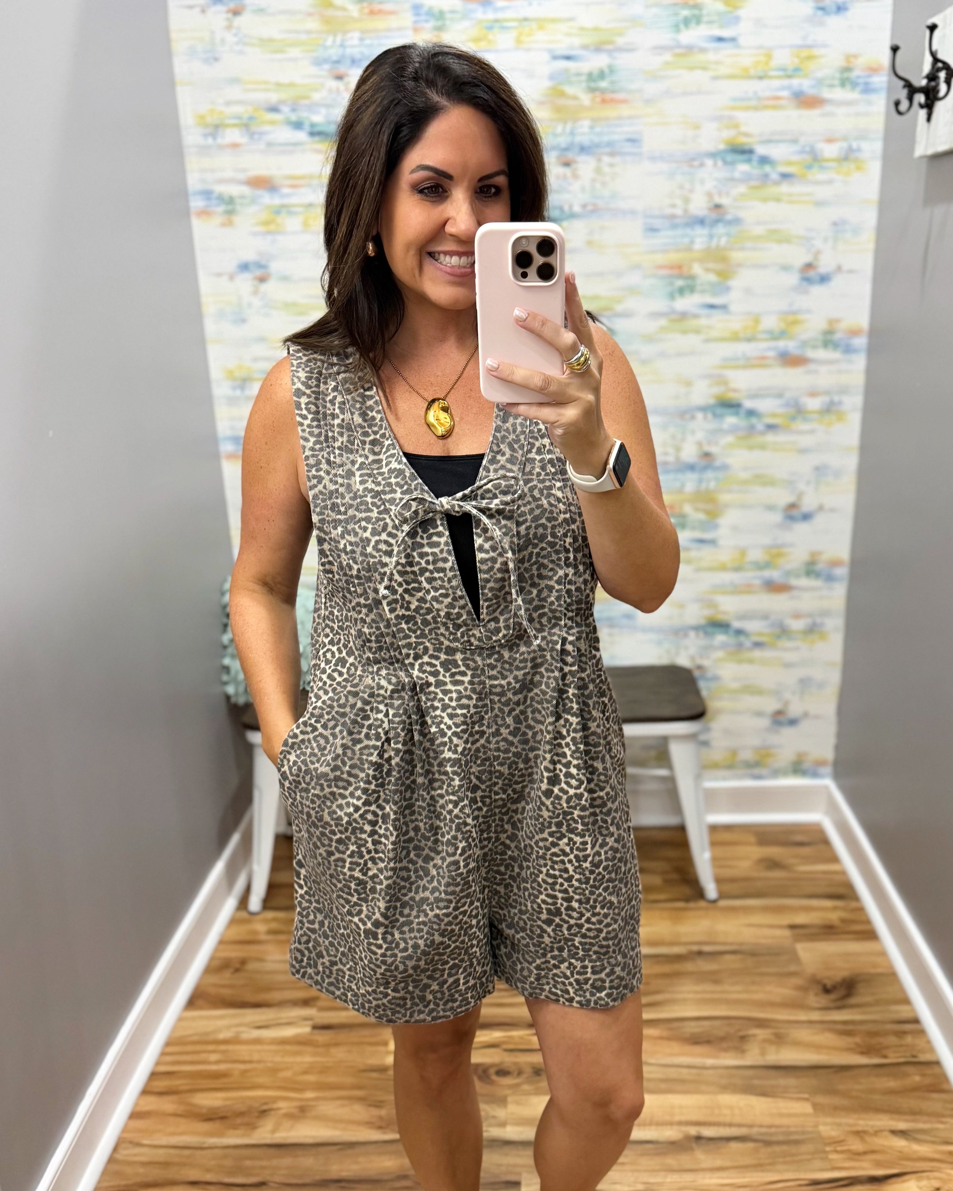A woman takes a mirror selfie wearing a sleeveless leopard-print romper with a tie at the neckline. The background shows a gray wall, a patterned wallpaper, a wooden floor, and a small white bench.