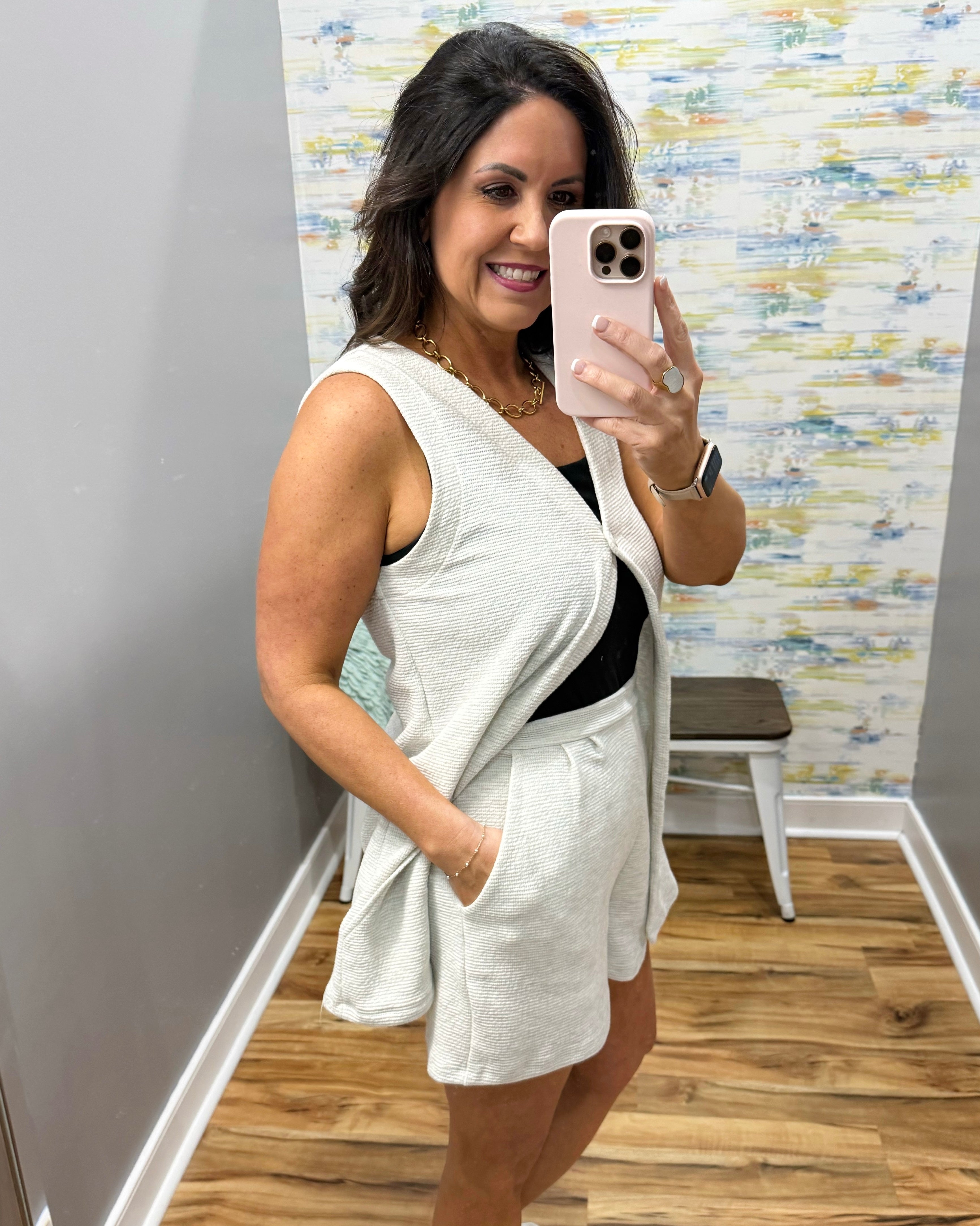 Woman taking a mirror selfie wearing a white sleeveless top and shorts in a room with a textured wall.