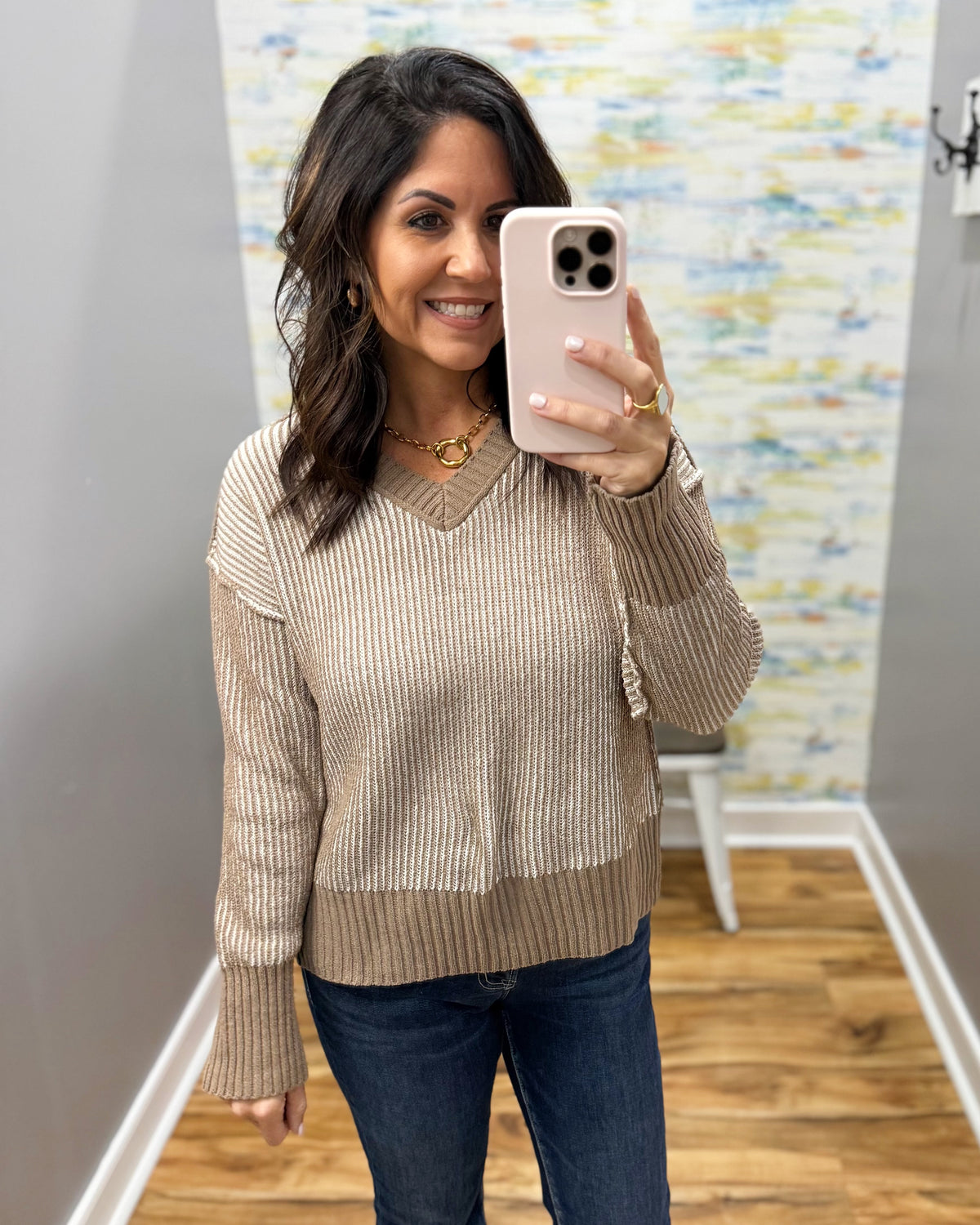 A woman takes a mirror selfie wearing a beige and brown ribbed knit sweater with dark blue jeans, holding a light pink smartphone.