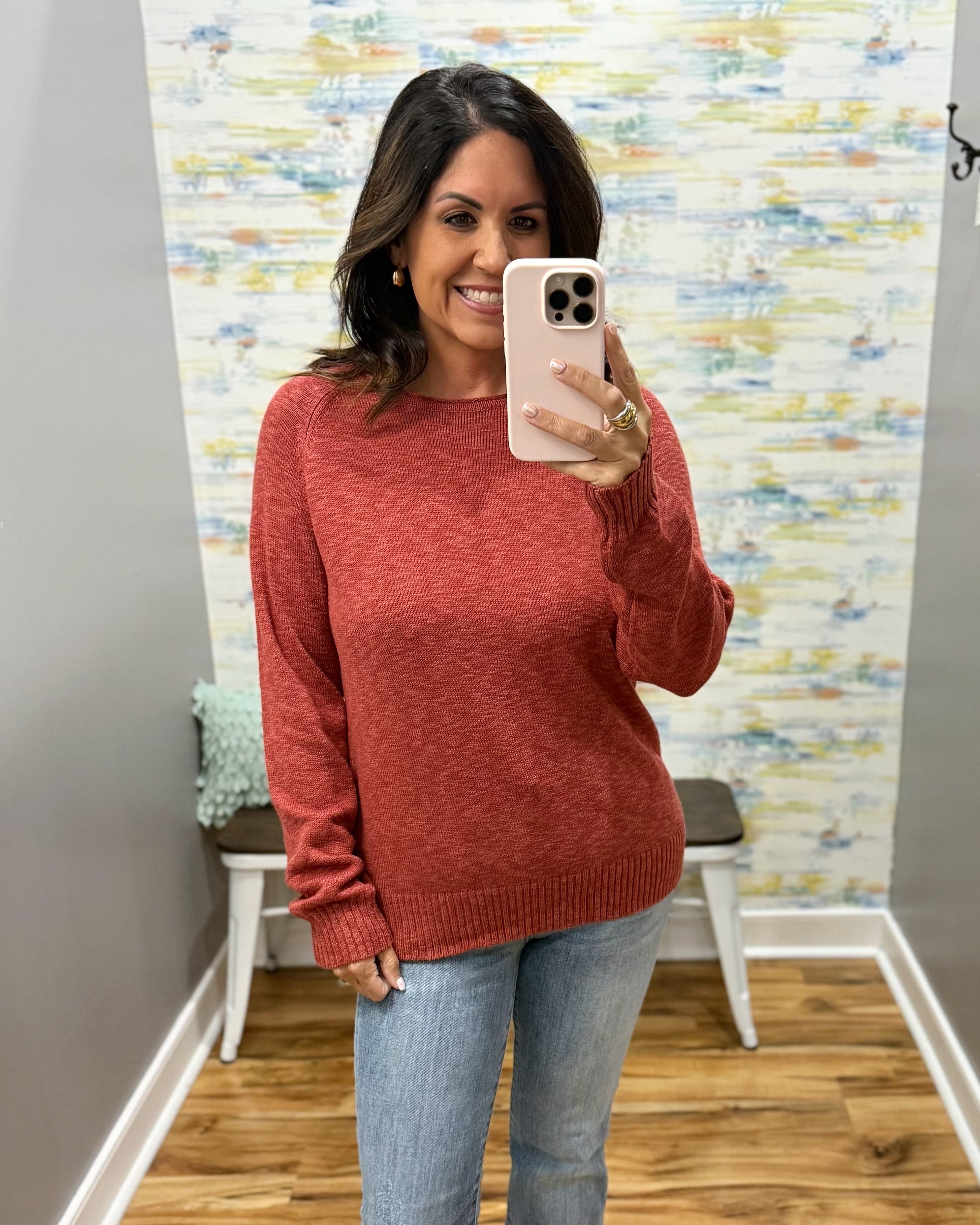 A woman takes a mirror selfie wearing a heathered rust-red long-sleeve knit sweater and light-wash blue jeans in a home fitting room with patterned wallpaper and wood floors.