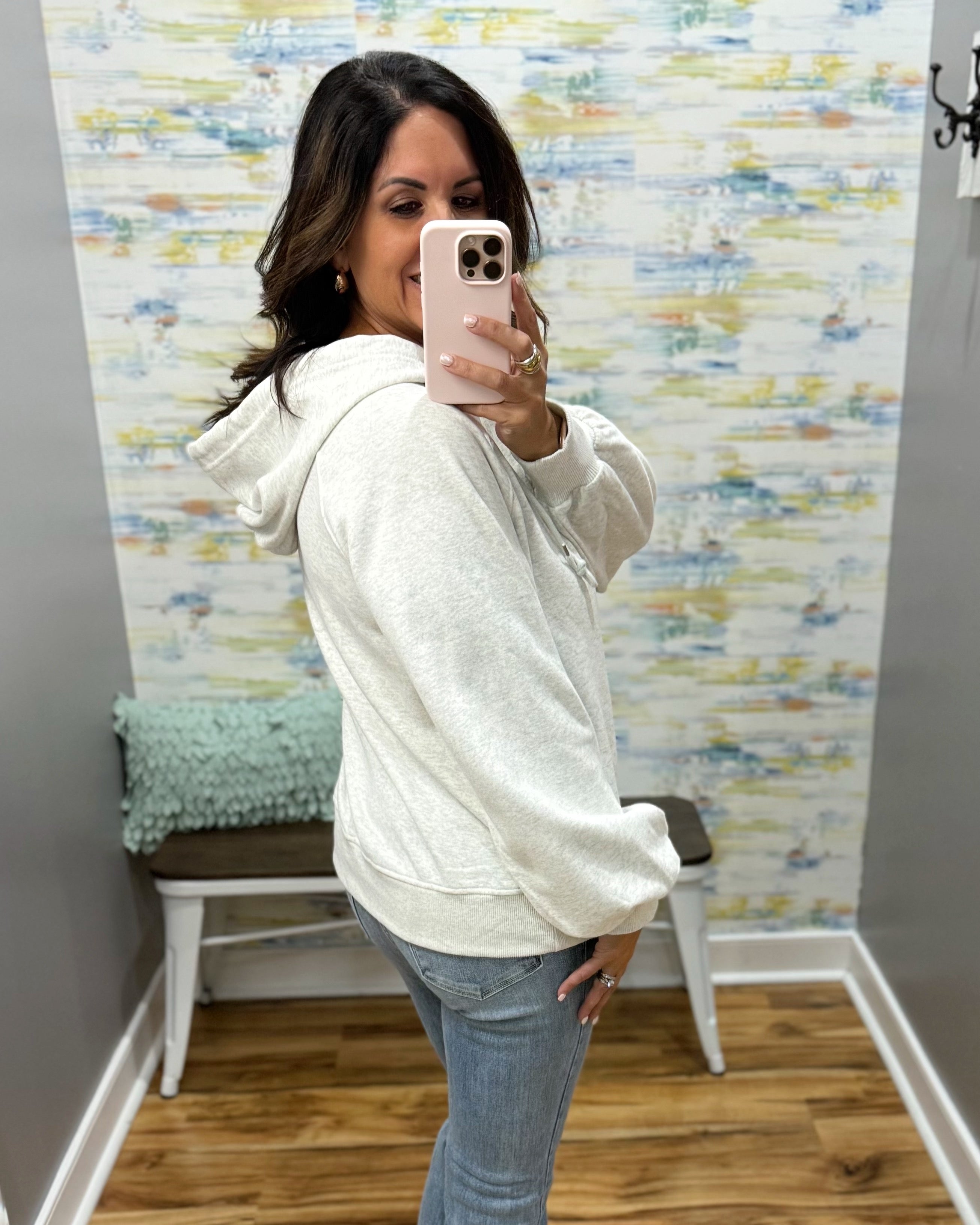 A woman takes a mirror selfie in a fitting room wearing a light heather gray hoodie and blue jeans, holding a light pink smartphone.