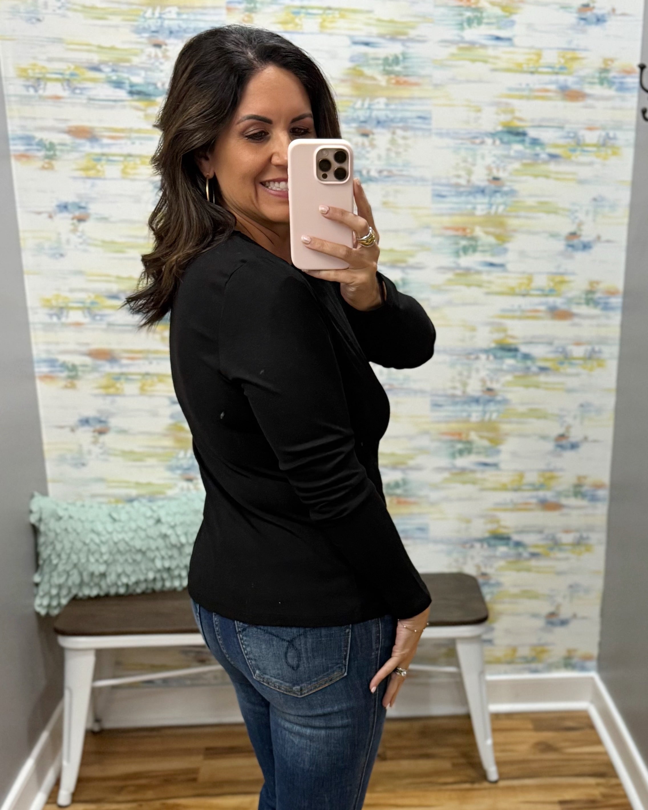 A woman takes a mirror selfie wearing a black long-sleeve top and blue jeans in front of a patterned wallpaper wall with a bench in the background.