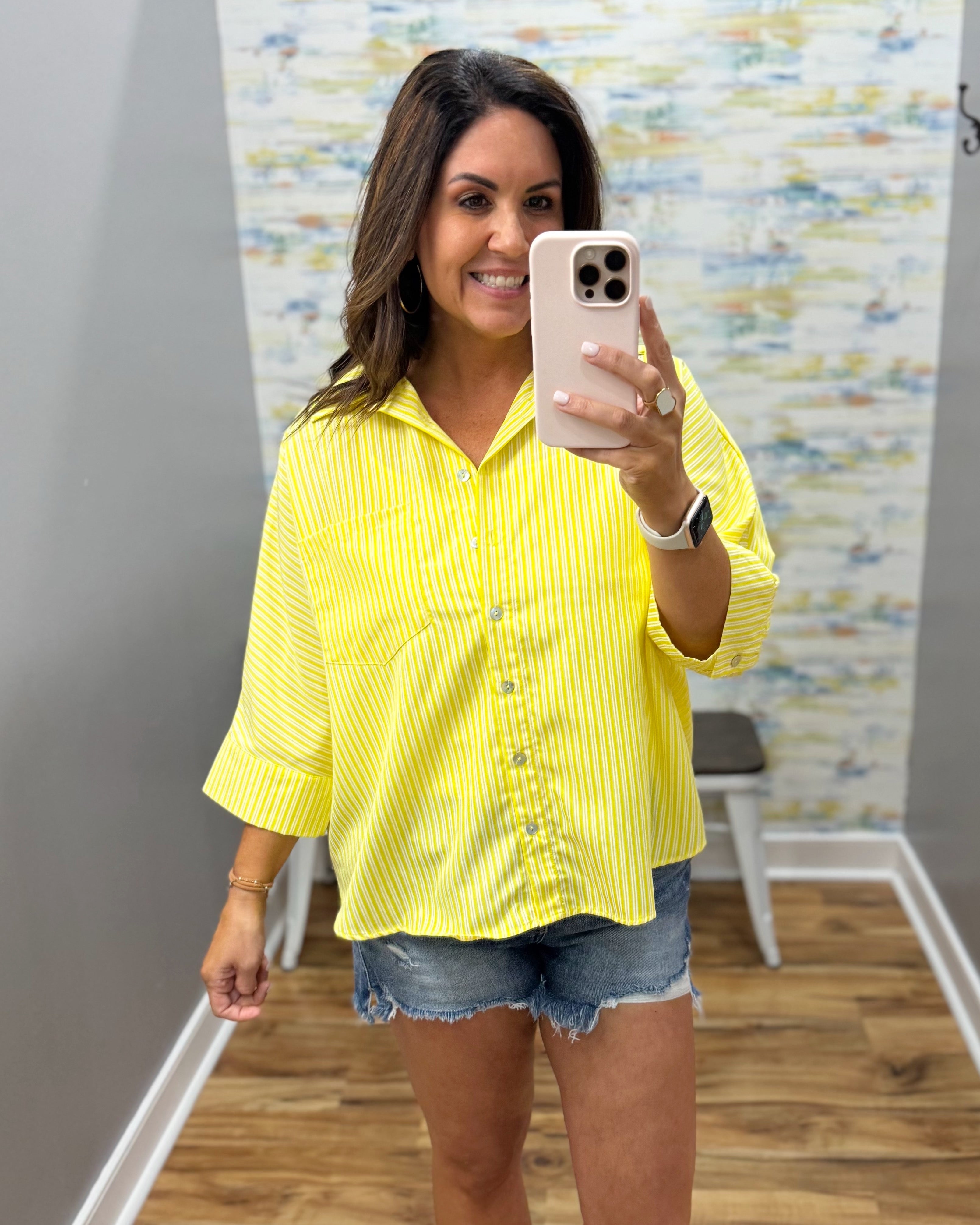 Woman taking a mirror selfie wearing a yellow striped shirt and denim shorts in a room with a colorful wall.