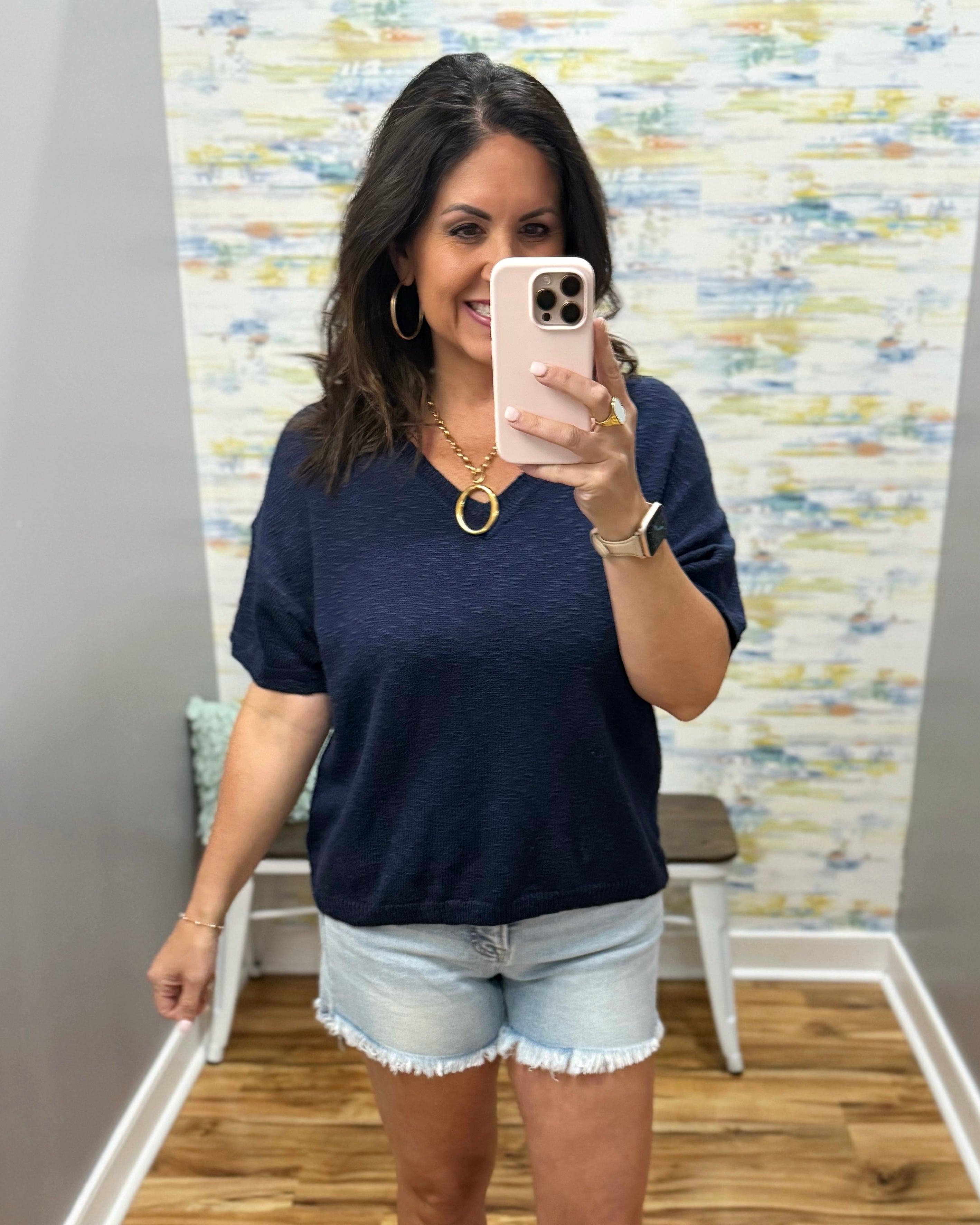 Woman taking a mirror selfie wearing a navy blue top and denim shorts in a room with colorful wall art.
