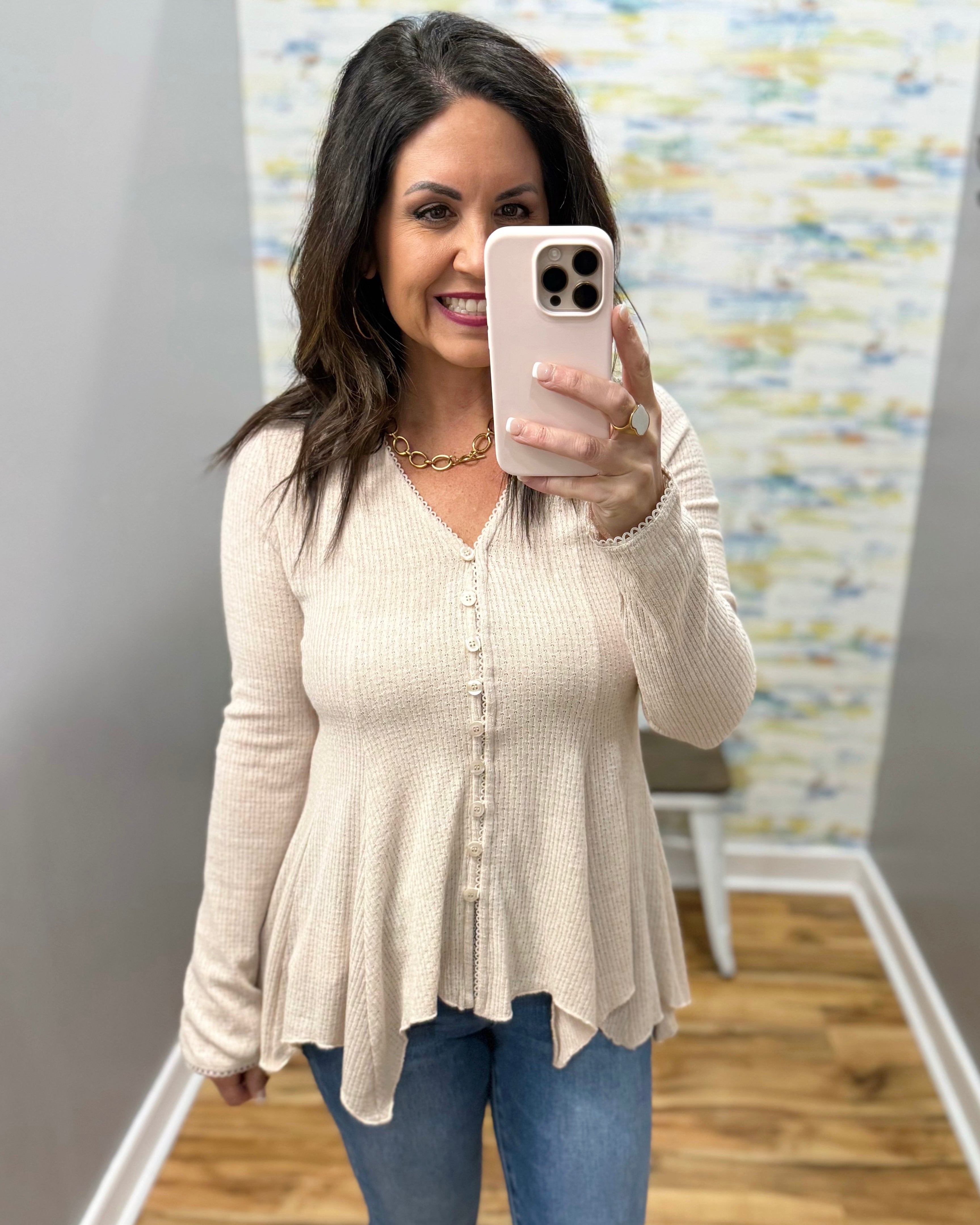 Woman taking a mirror selfie wearing a beige cardigan and blue jeans in a room with a textured wall.