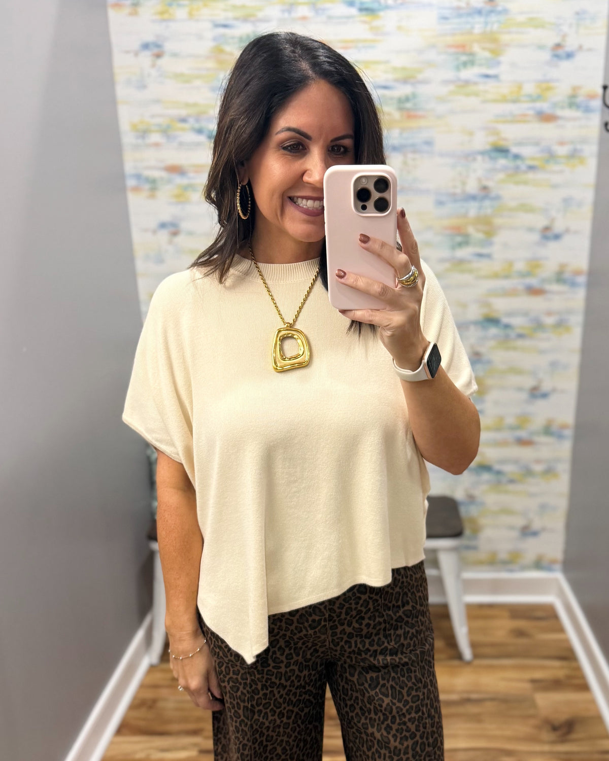 A woman taking a mirror selfie wearing a cream-colored short-sleeve top and leopard print pants, accessorized with a gold necklace and hoop earrings.