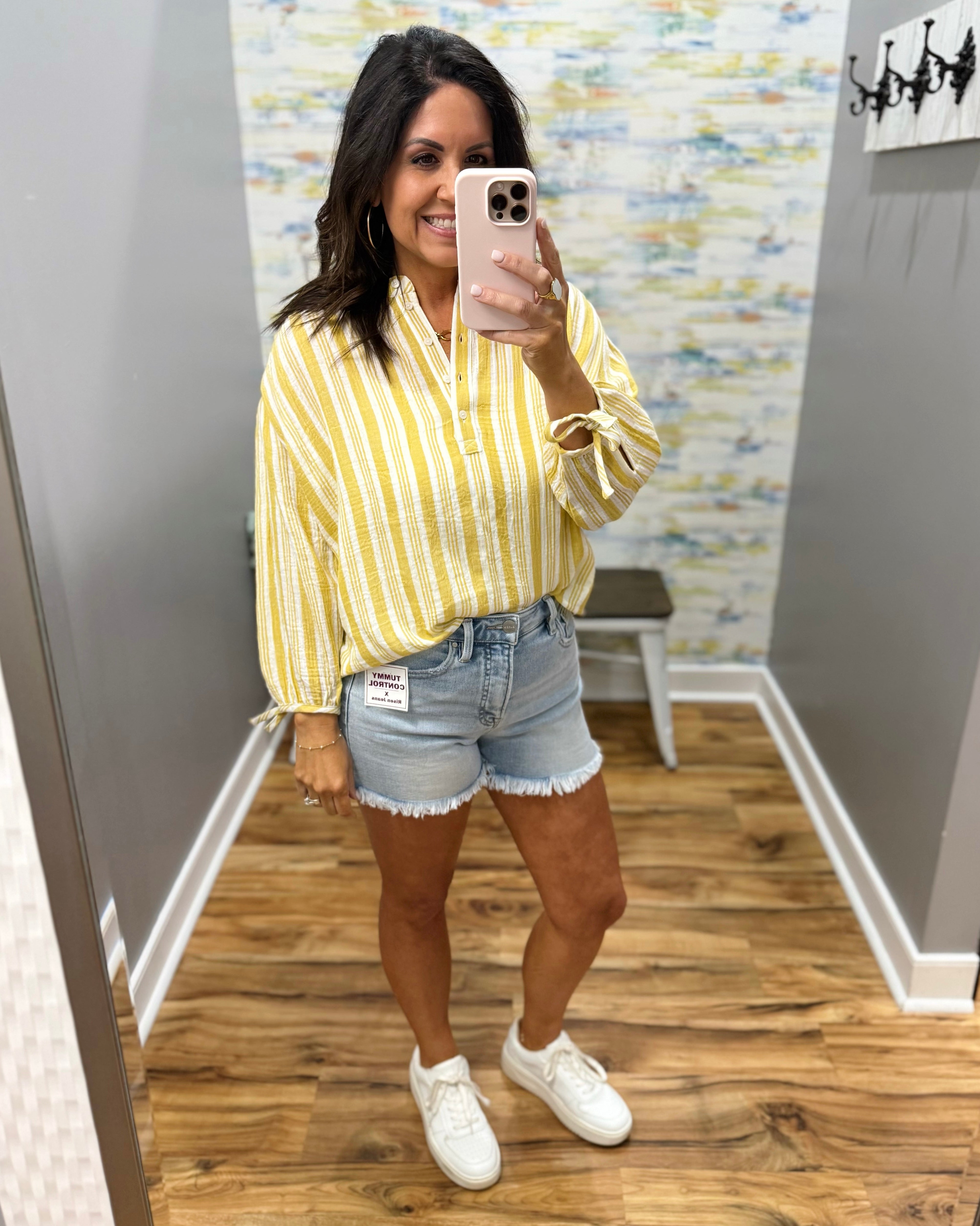 Woman taking a mirror selfie wearing a yellow striped shirt, denim shorts, and white sneakers in a room with a patterned wall.