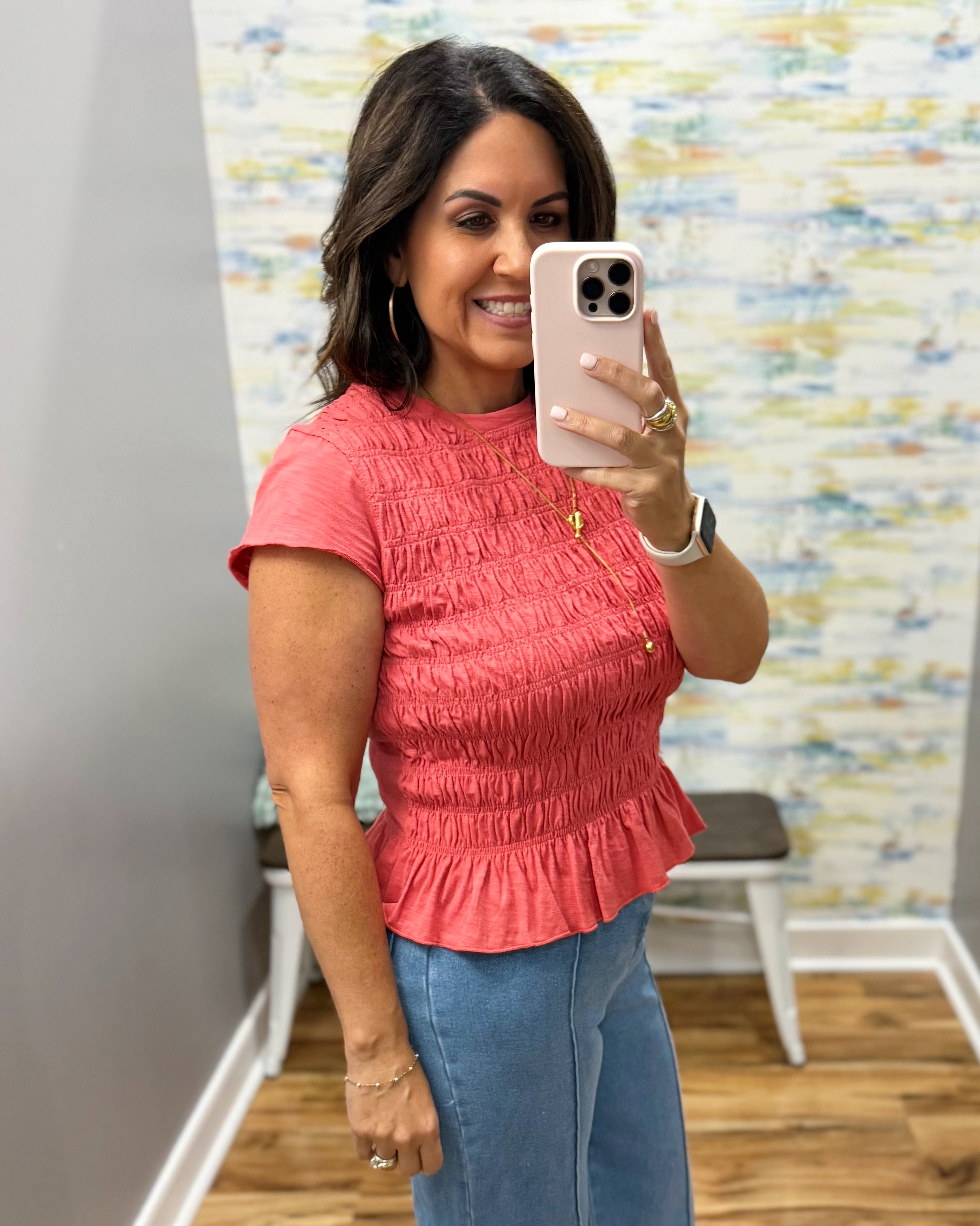 A woman takes a mirror selfie wearing a coral pink short-sleeve smocked peplum top and blue jeans in a fitting room.