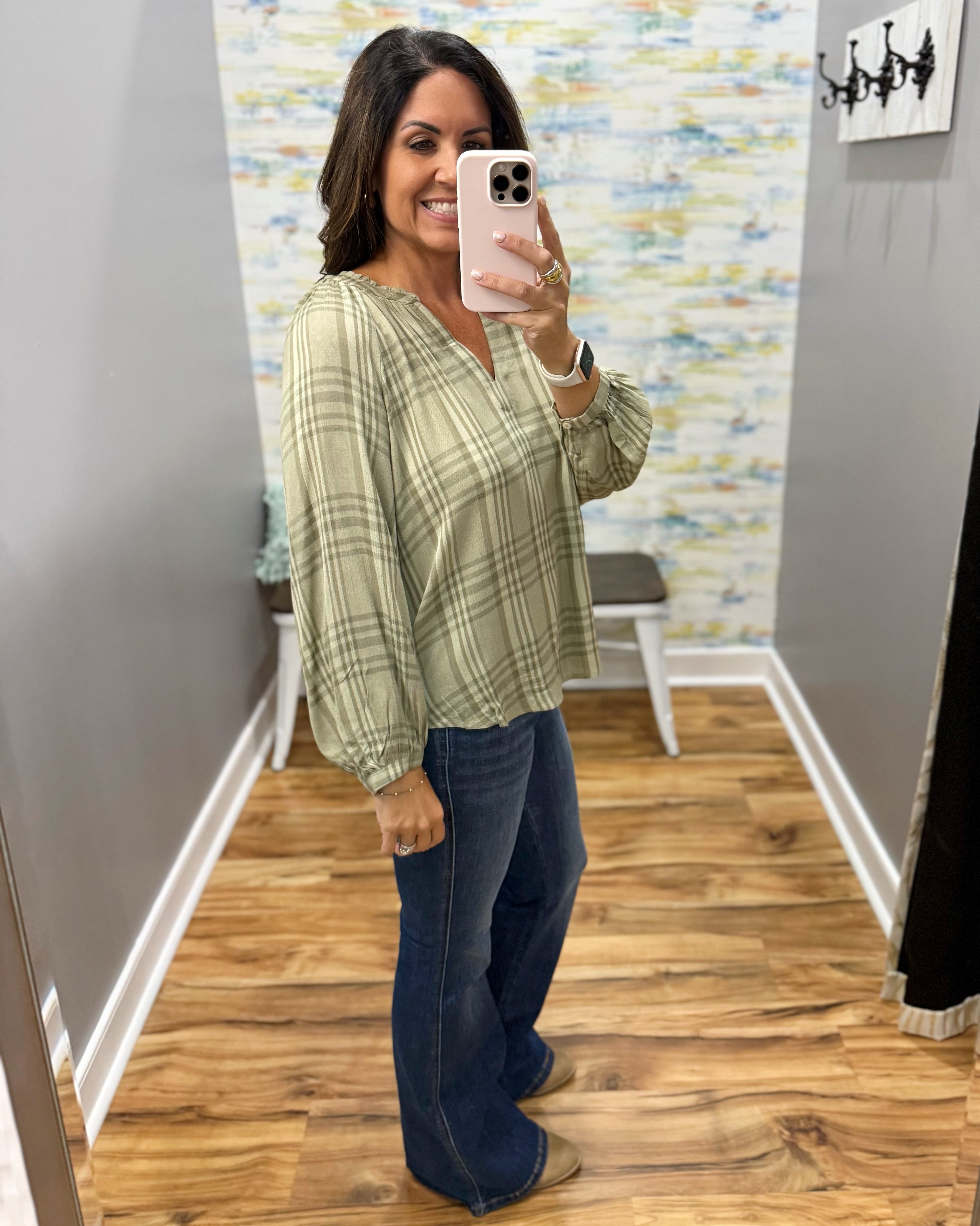 A woman takes a mirror selfie in a fitting room wearing a sage green and beige plaid long-sleeve blouse with dark blue flare jeans.