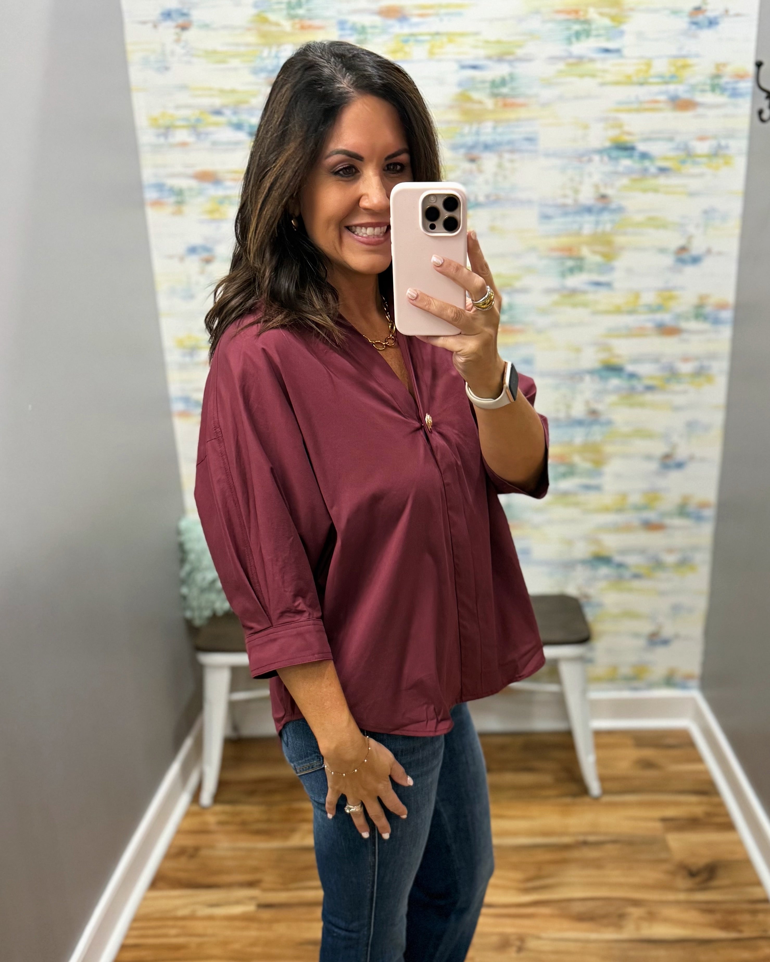 A woman takes a mirror selfie wearing a loose-fitting, three-quarter sleeve button-up blouse in a deep burgundy or maroon color, paired with blue jeans.