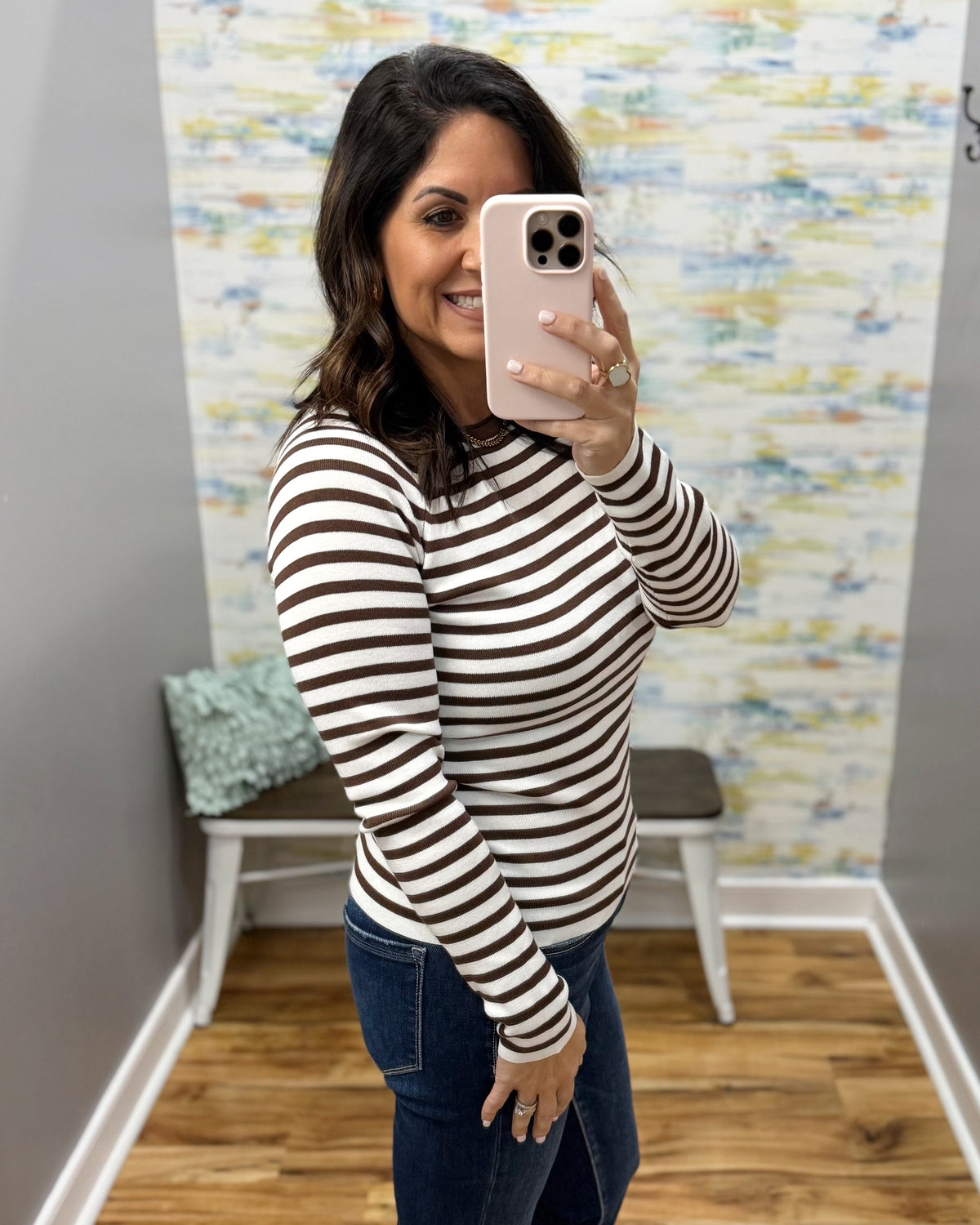 A woman takes a mirror selfie wearing a brown and white horizontally striped long-sleeve top and dark blue jeans in a fitting room with a patterned wall and wood floor.