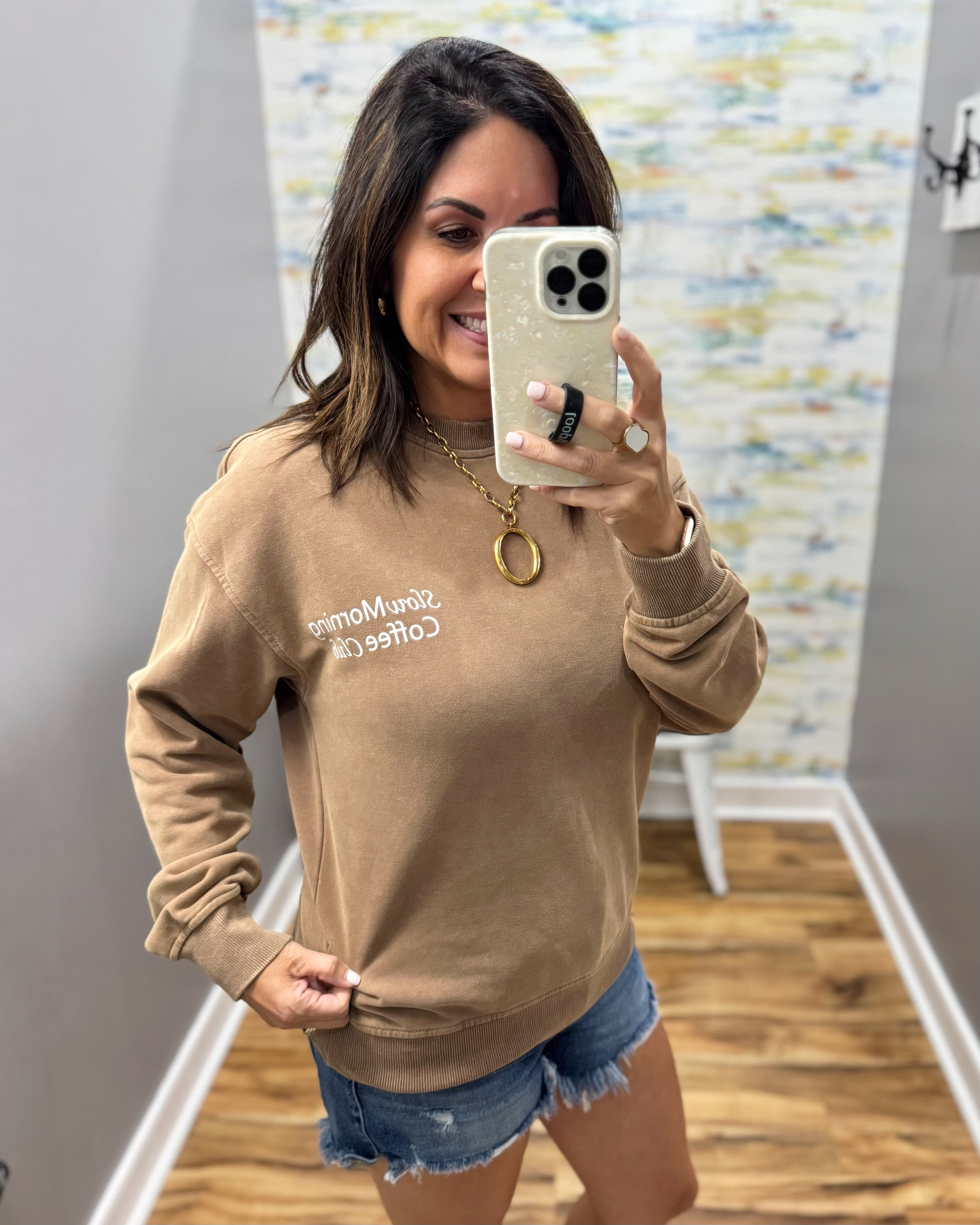 A woman takes a mirror selfie wearing a tan crewneck sweatshirt with white embroidered text that reads 'She's My Man Coffee' and denim shorts in a home setting.