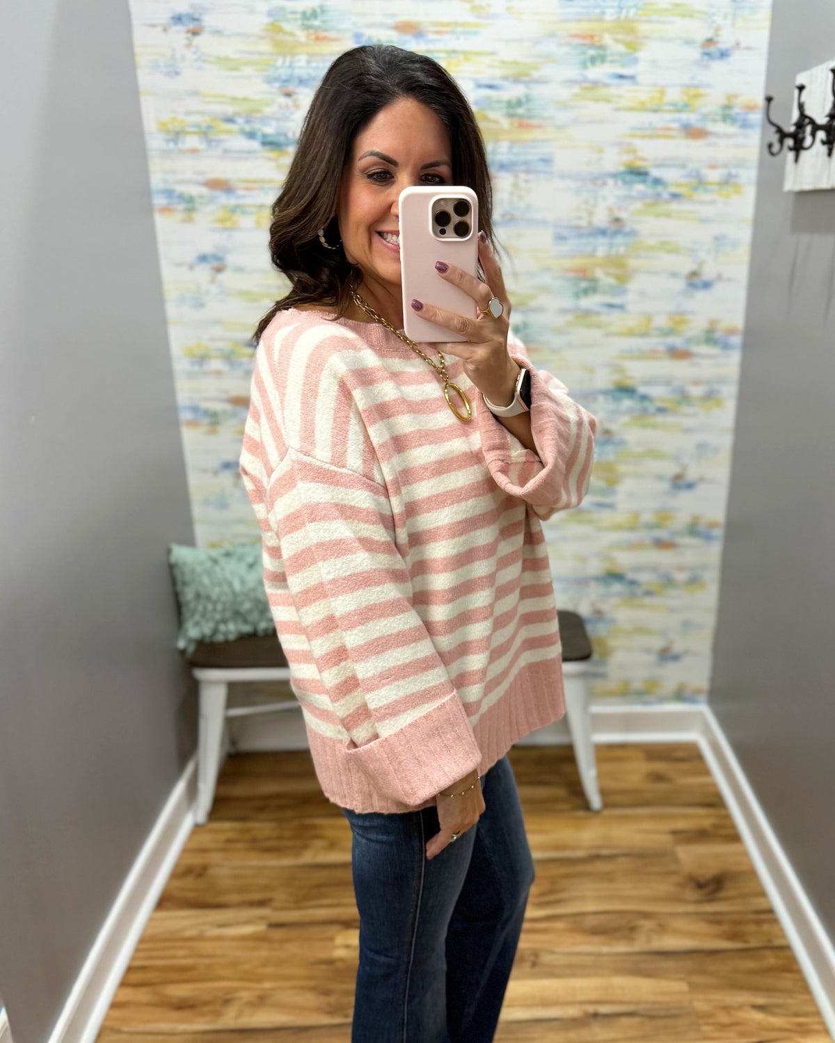 A woman takes a mirror selfie wearing a pink and cream striped knit sweater, blue jeans, and gold jewelry in a home hallway.