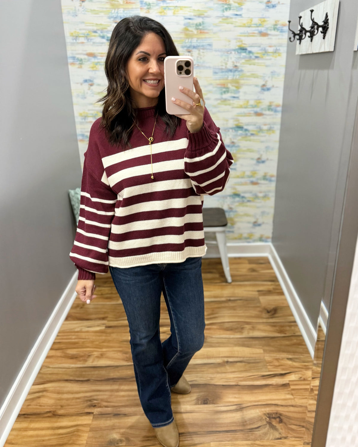 A woman takes a mirror selfie wearing a burgundy and cream striped knit sweater, blue jeans, and tan boots in a home hallway.