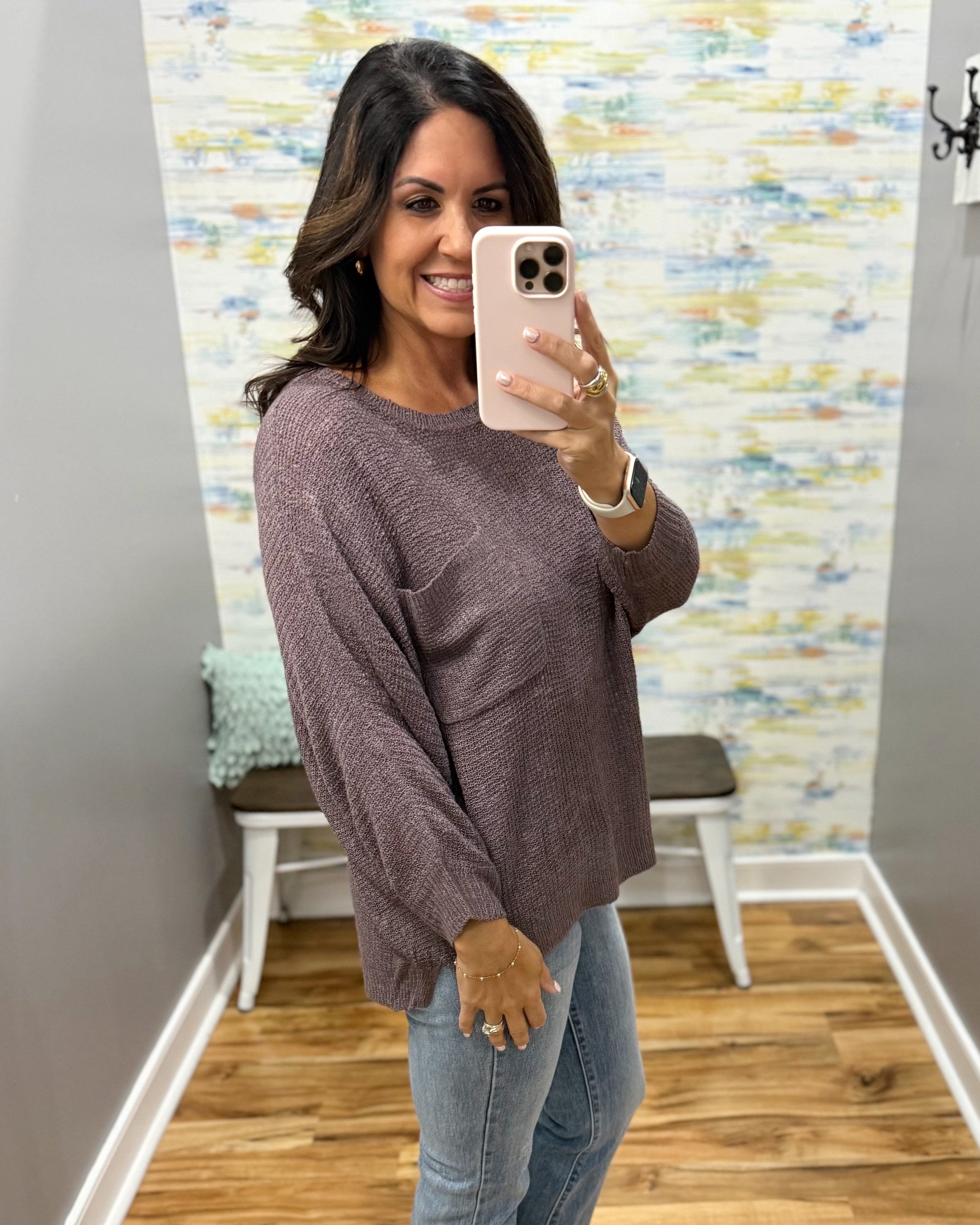A woman takes a mirror selfie wearing a textured, long-sleeve knit sweater in a muted mauve-brown color, paired with light-wash blue jeans.