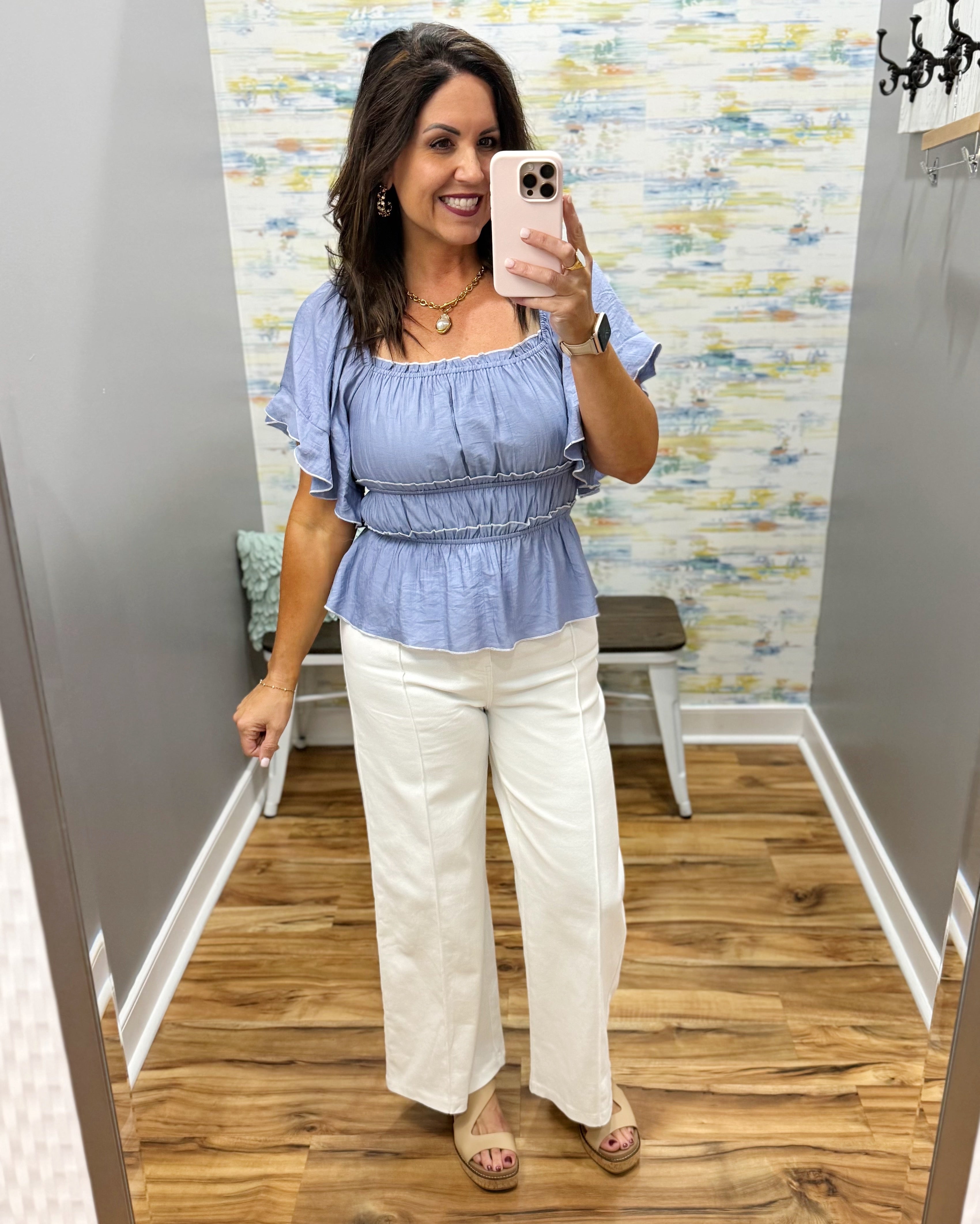 Woman taking a mirror selfie wearing a blue top and white pants in a store.