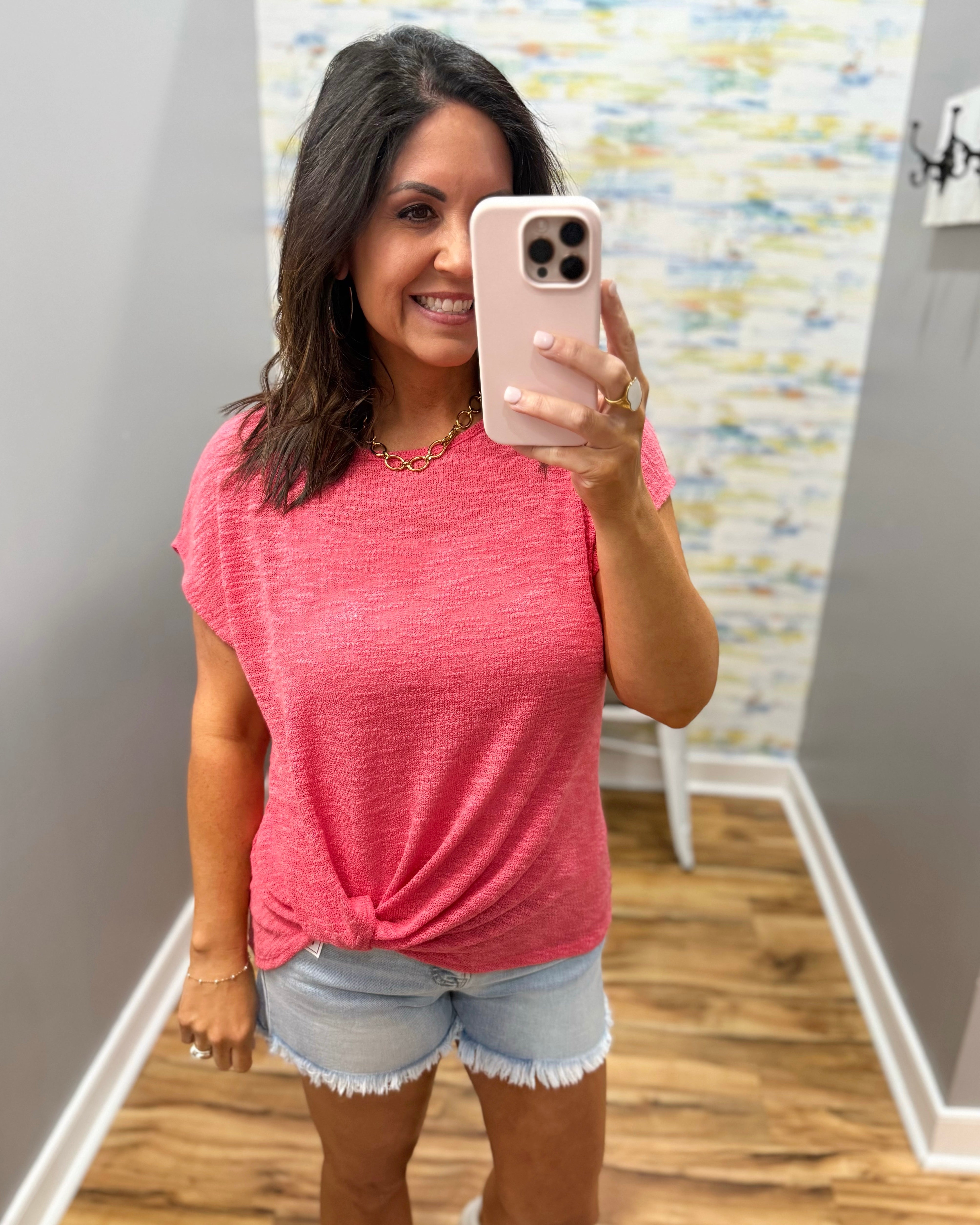Woman taking a mirror selfie wearing a pink top and denim shorts in a room with a colorful wall.