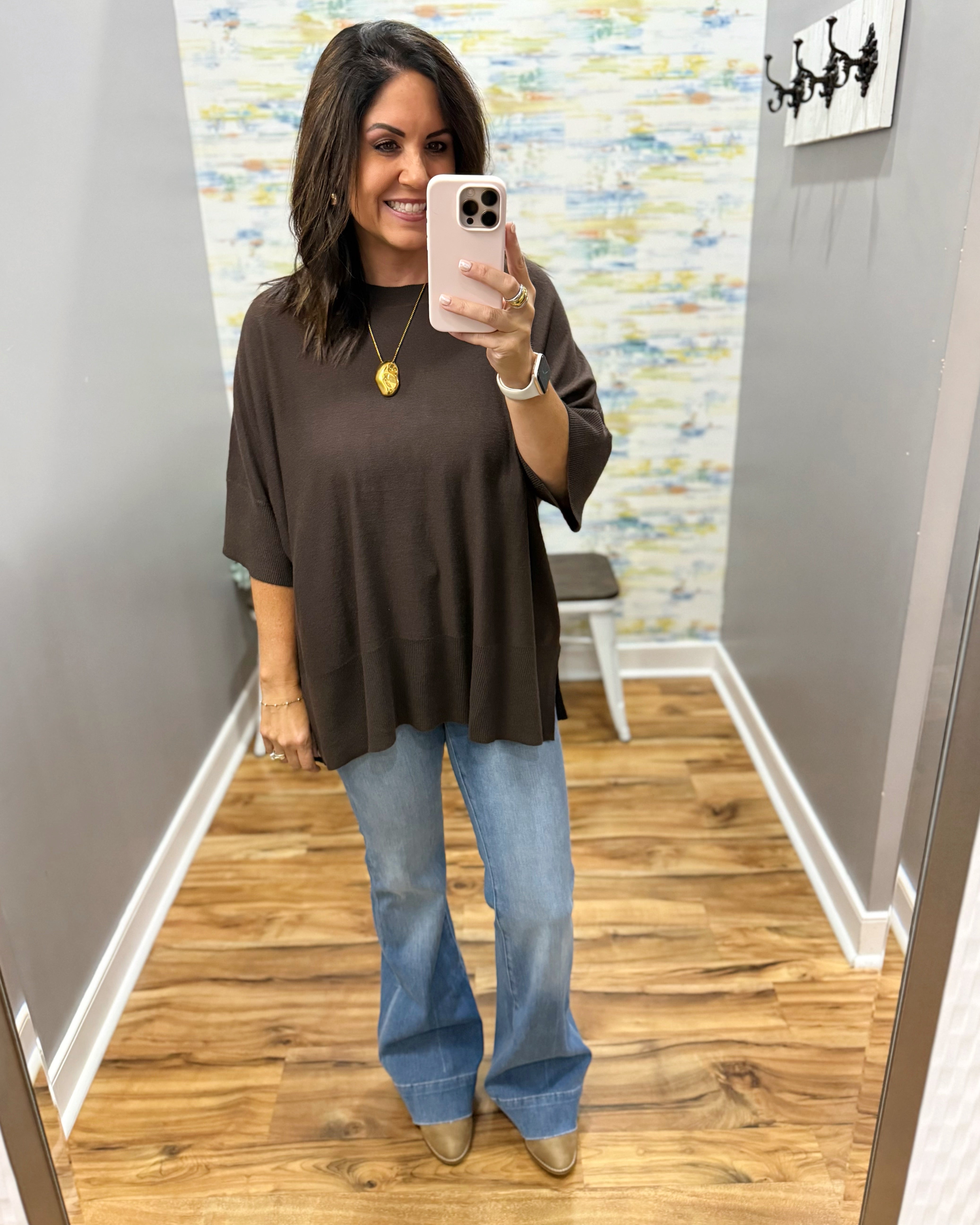 A woman takes a mirror selfie in a fitting room wearing a dark brown oversized short-sleeve top, light blue wide-leg jeans, and beige pointed-toe flats.