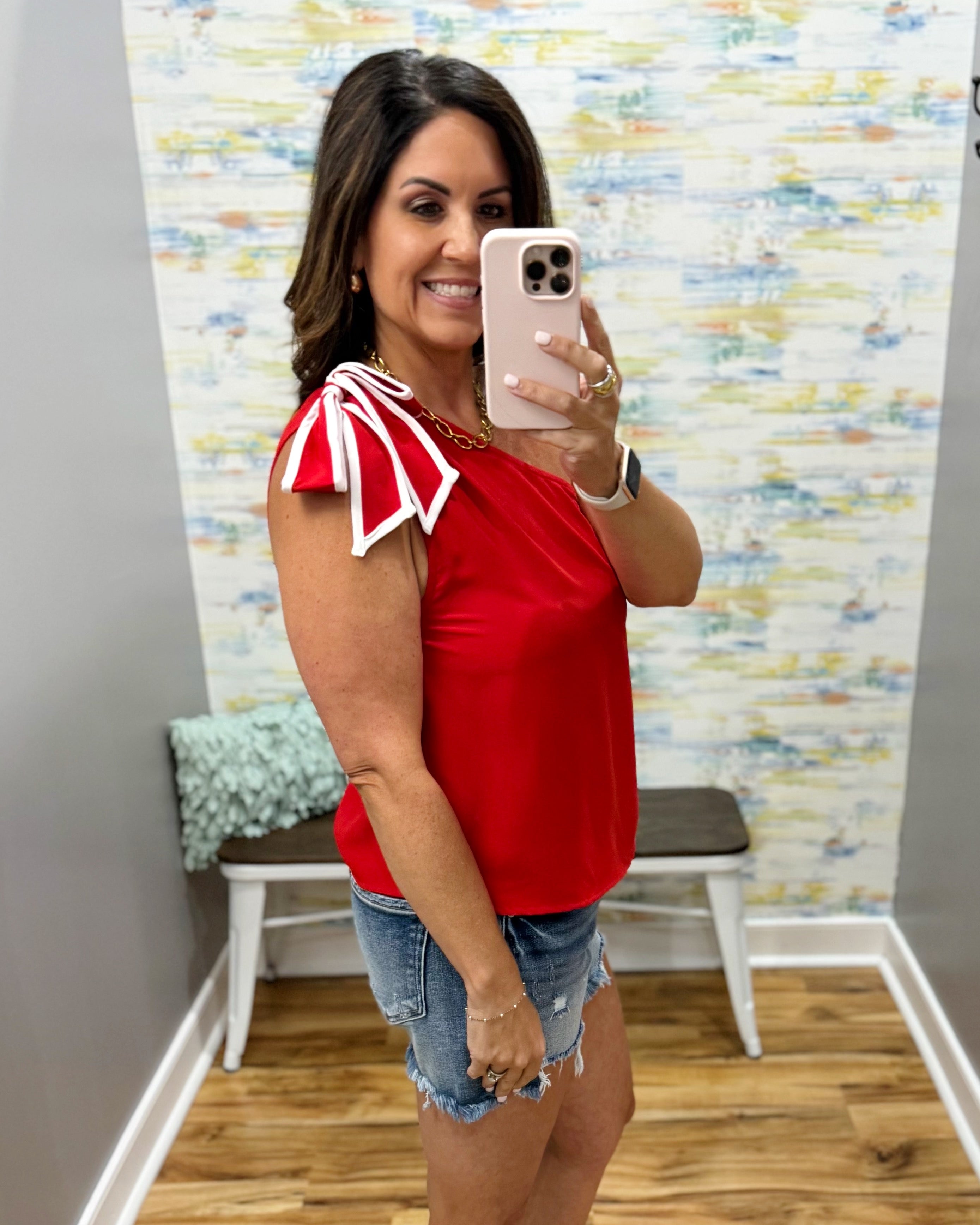 Woman taking a mirror selfie wearing a red top with white accents and denim shorts in a room with a colorful wall.