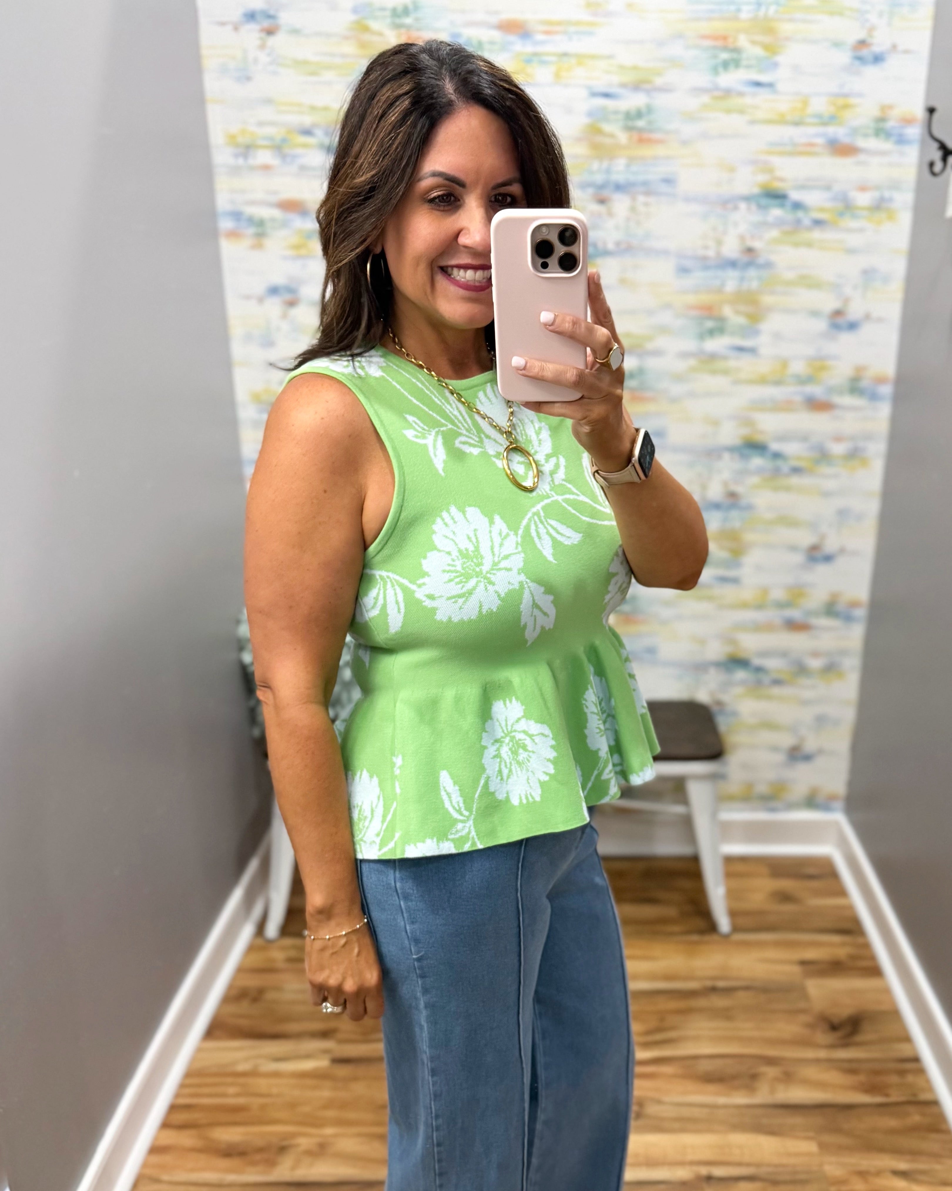 Woman taking a mirror selfie wearing a green floral top and blue jeans in a room with a colorful wall.