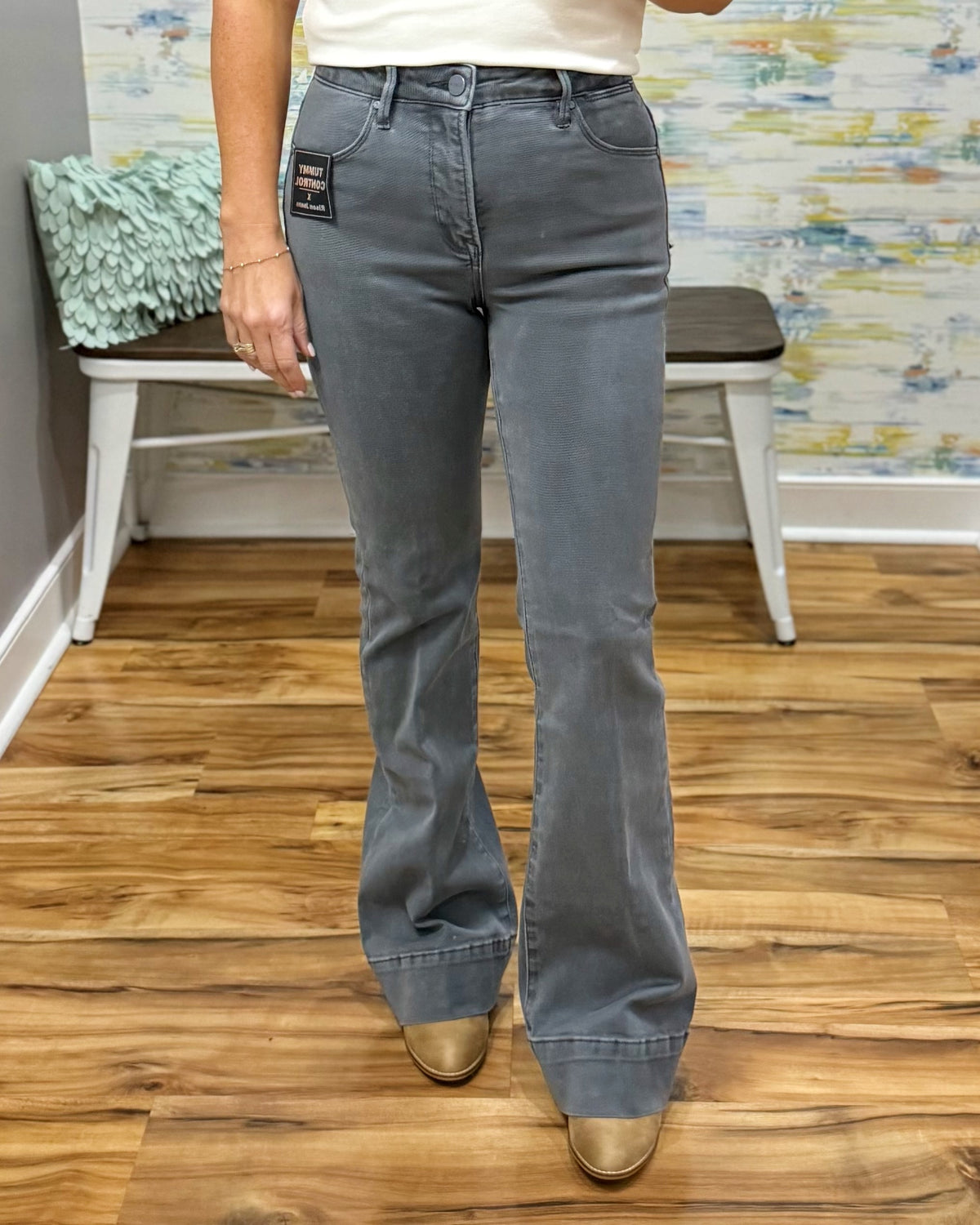 A person wearing gray flare jeans with a white top and tan boots, standing on a wood floor in front of a bench and abstract wall art.