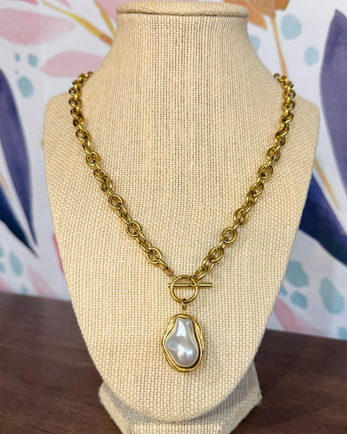 Chansuttpearls Pearl Drop necklace with a large irregular white pearl pendant, displayed on a beige fabric jewelry bust against a colorful floral background.