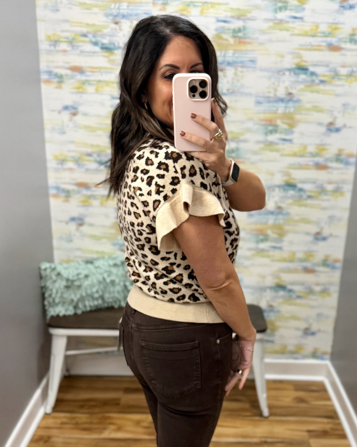 A woman takes a mirror selfie wearing a beige and black leopard print short-sleeve top with ruffled sleeves and dark brown pants, standing in an indoor room with a patterned wallpaper background.
