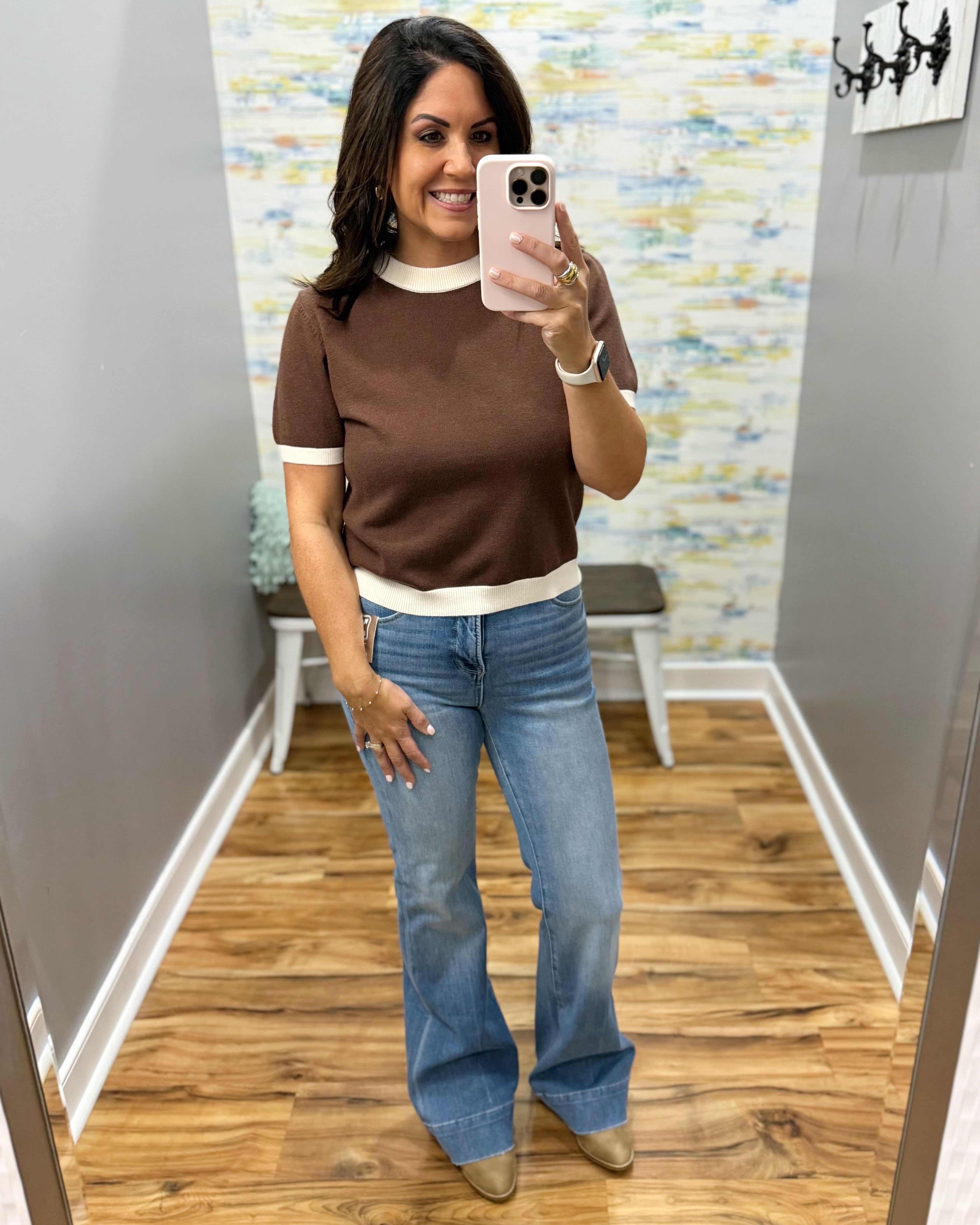 A woman takes a mirror selfie in a fitting room wearing a brown short-sleeve knit top with cream ribbed trim and light blue flare jeans.