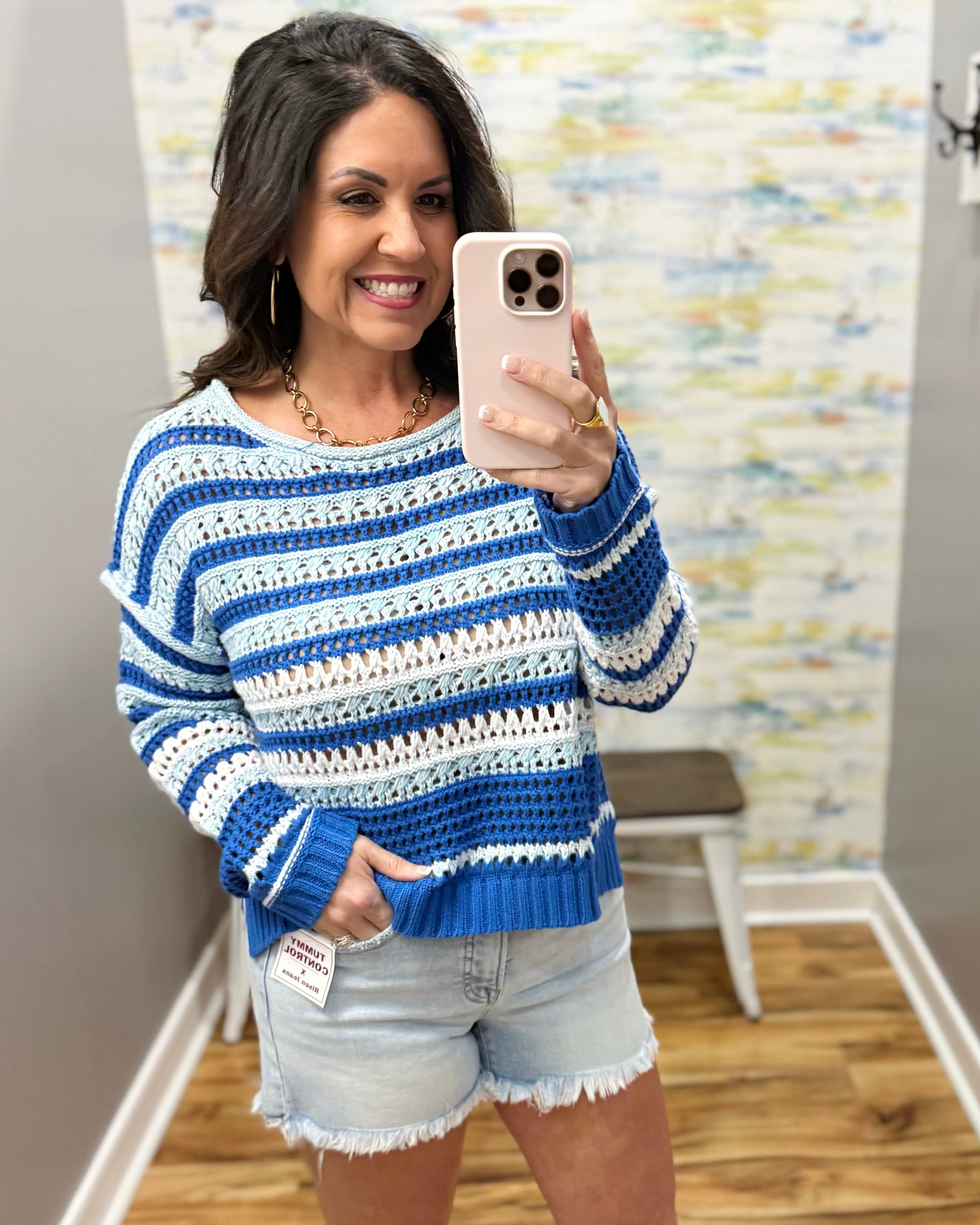 Woman wearing a blue and white striped sweater and denim shorts, taking a mirror selfie.