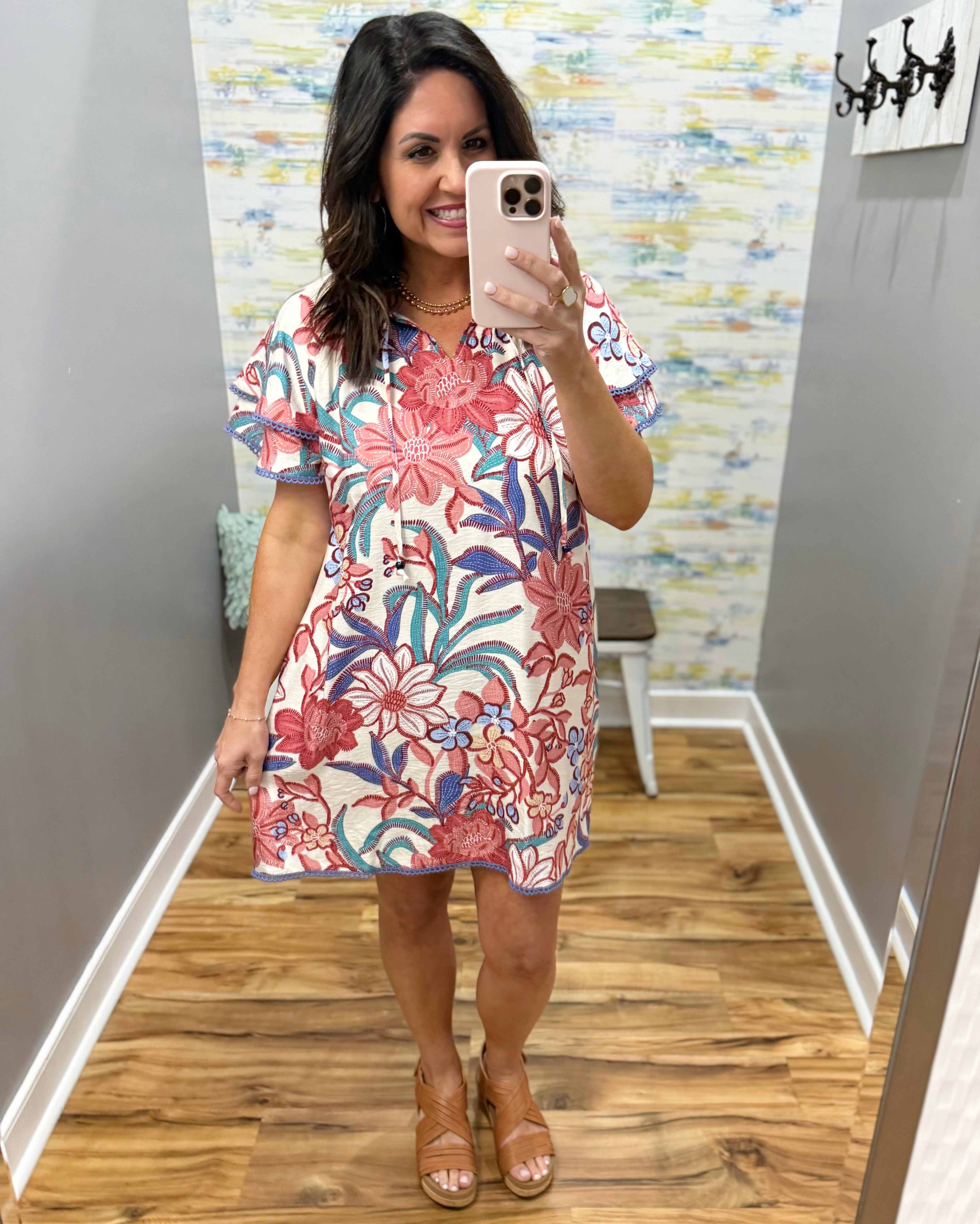 Woman taking a mirror selfie wearing a floral dress in a room with colorful wallpaper.