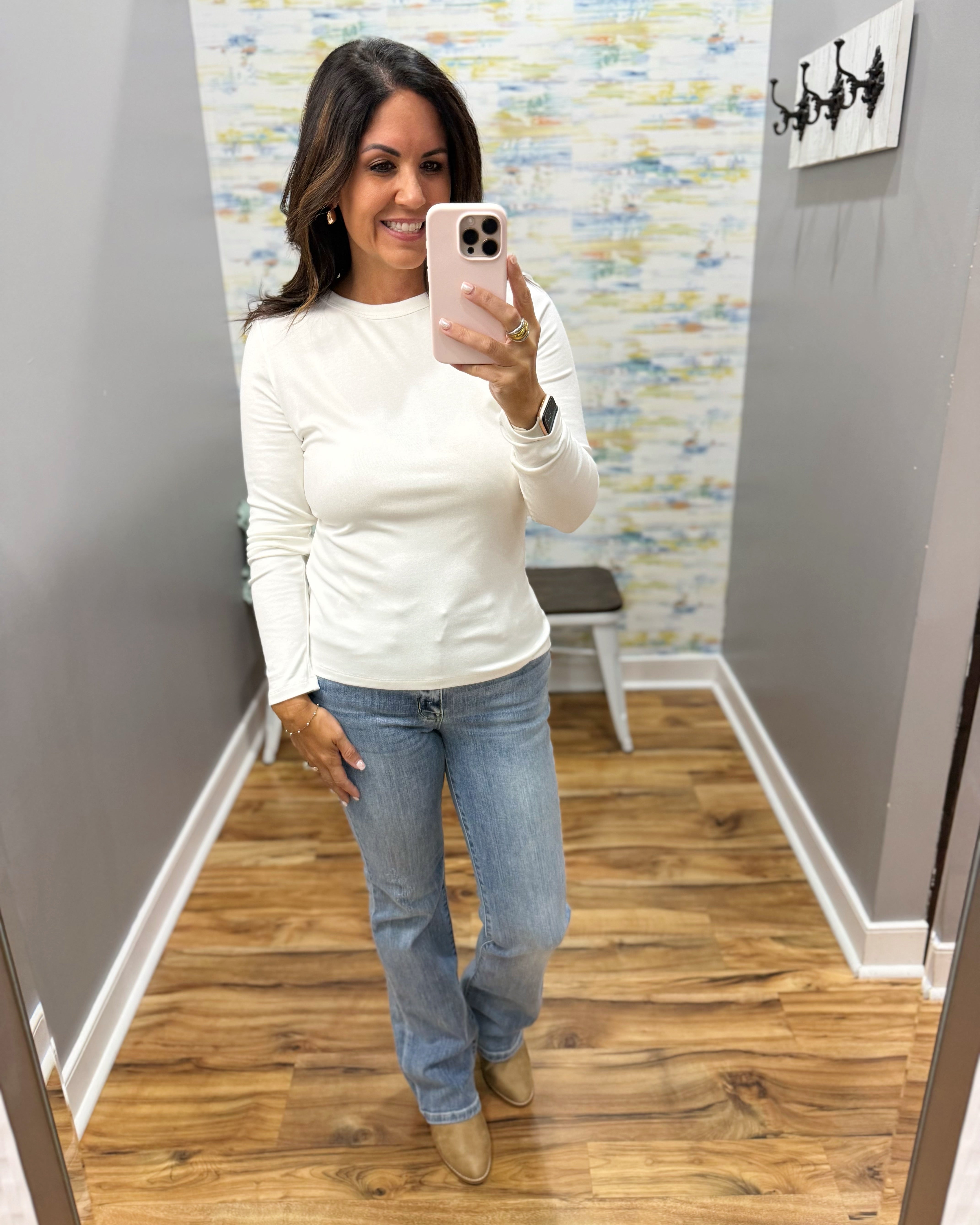 A woman takes a mirror selfie wearing a fitted white long-sleeve crewneck top, light-wash blue jeans, and tan ankle boots in a fitting room with wood floors and gray walls.
