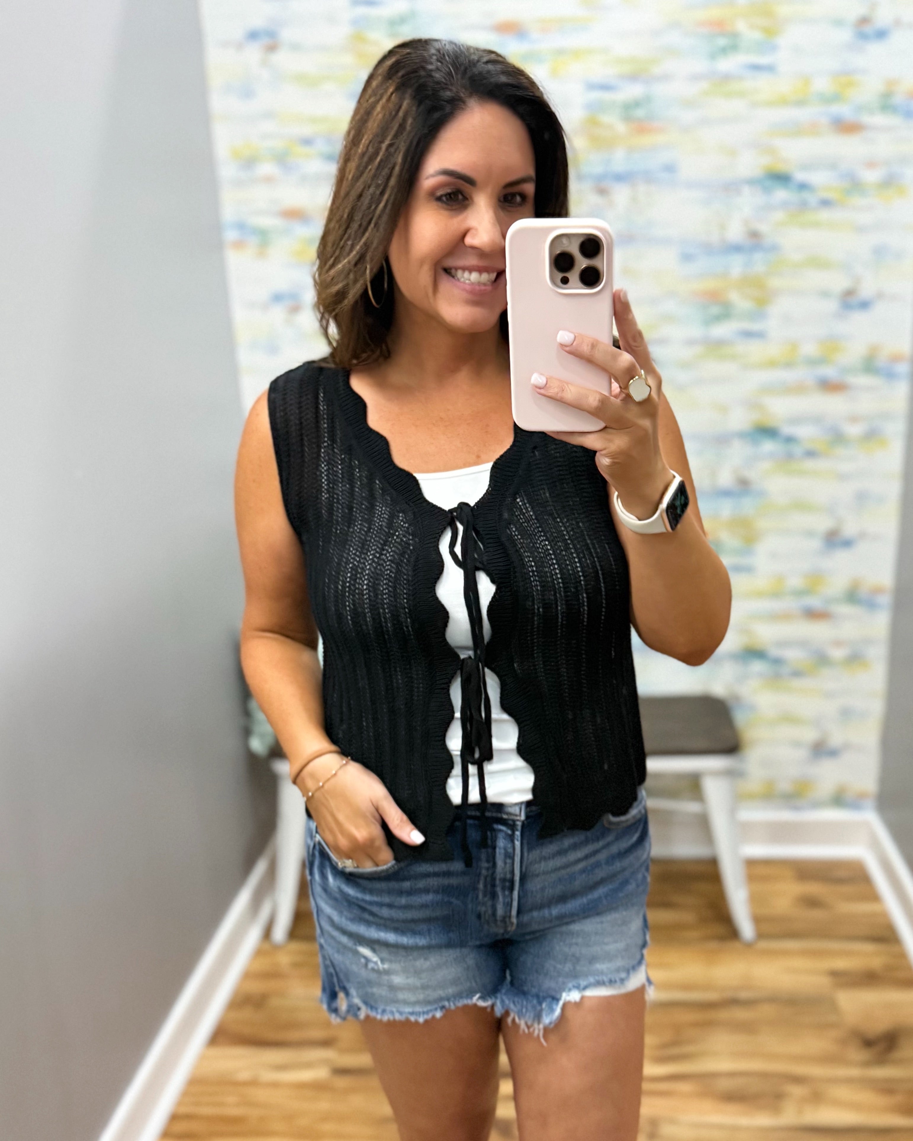 Woman taking a mirror selfie wearing a black sleeveless top and denim shorts in a room with a colorful wall.