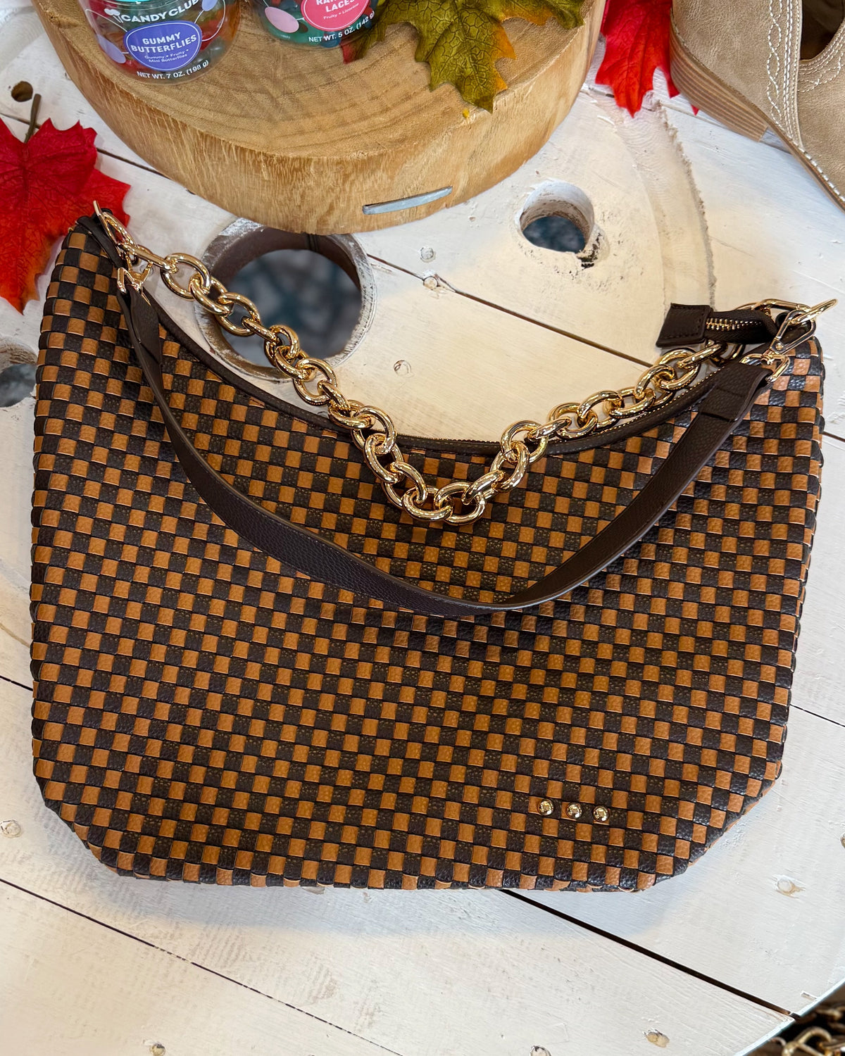 A brown and tan checkerboard woven handbag with a gold chain strap and dark brown leather shoulder strap, displayed on a white wooden surface with autumn leaves and other decorative items in the background.