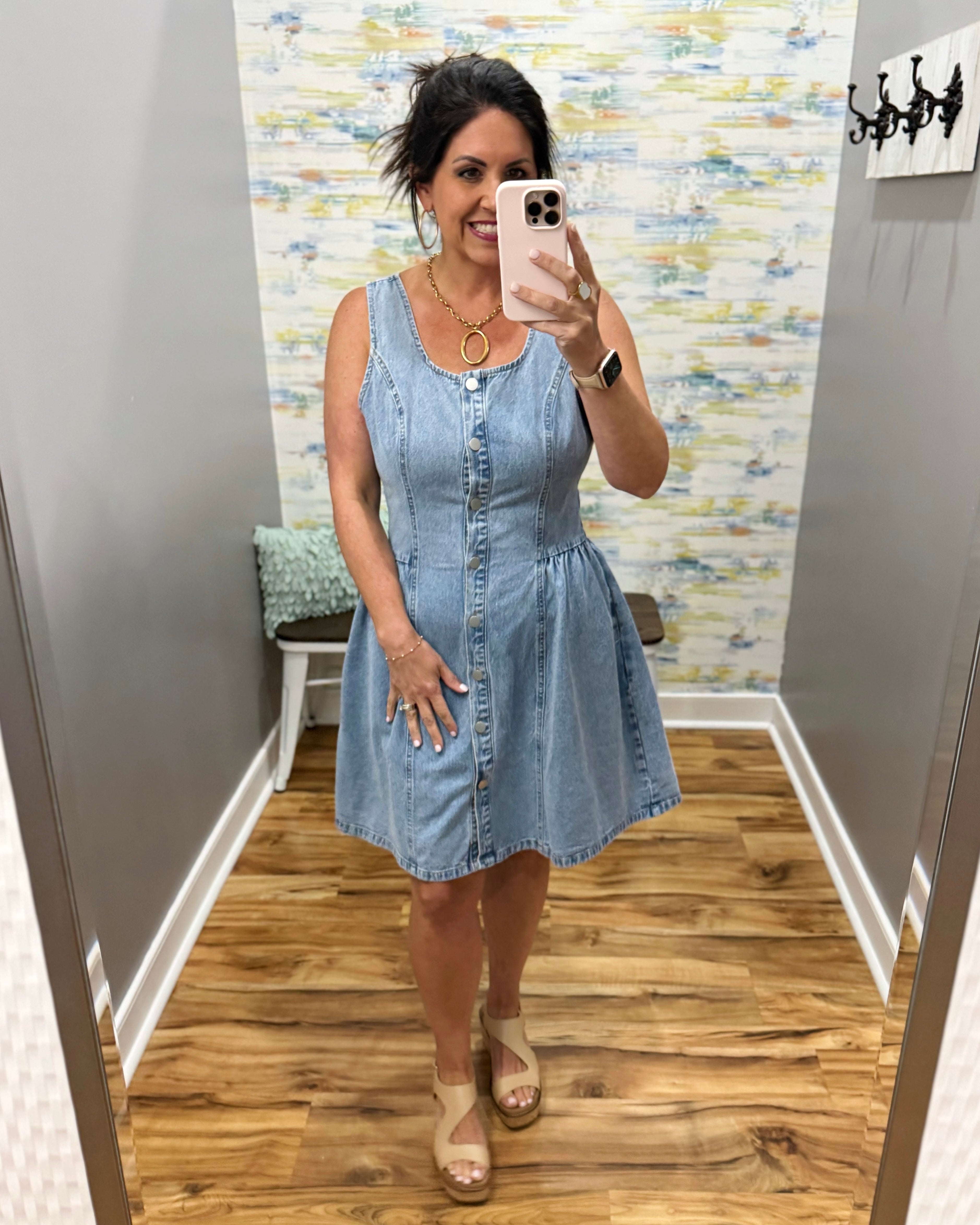 Woman taking a mirror selfie wearing a light blue denim dress in a room with a patterned wall.