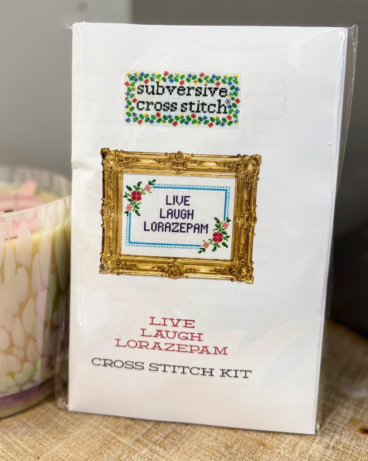 A packaged cross stitch kit labeled 'subversive cross stitch' and 'LIVE LAUGH LORAZEPAM' in a white box with a gold frame illustration and floral accents, photographed on a tabletop next to a candle.