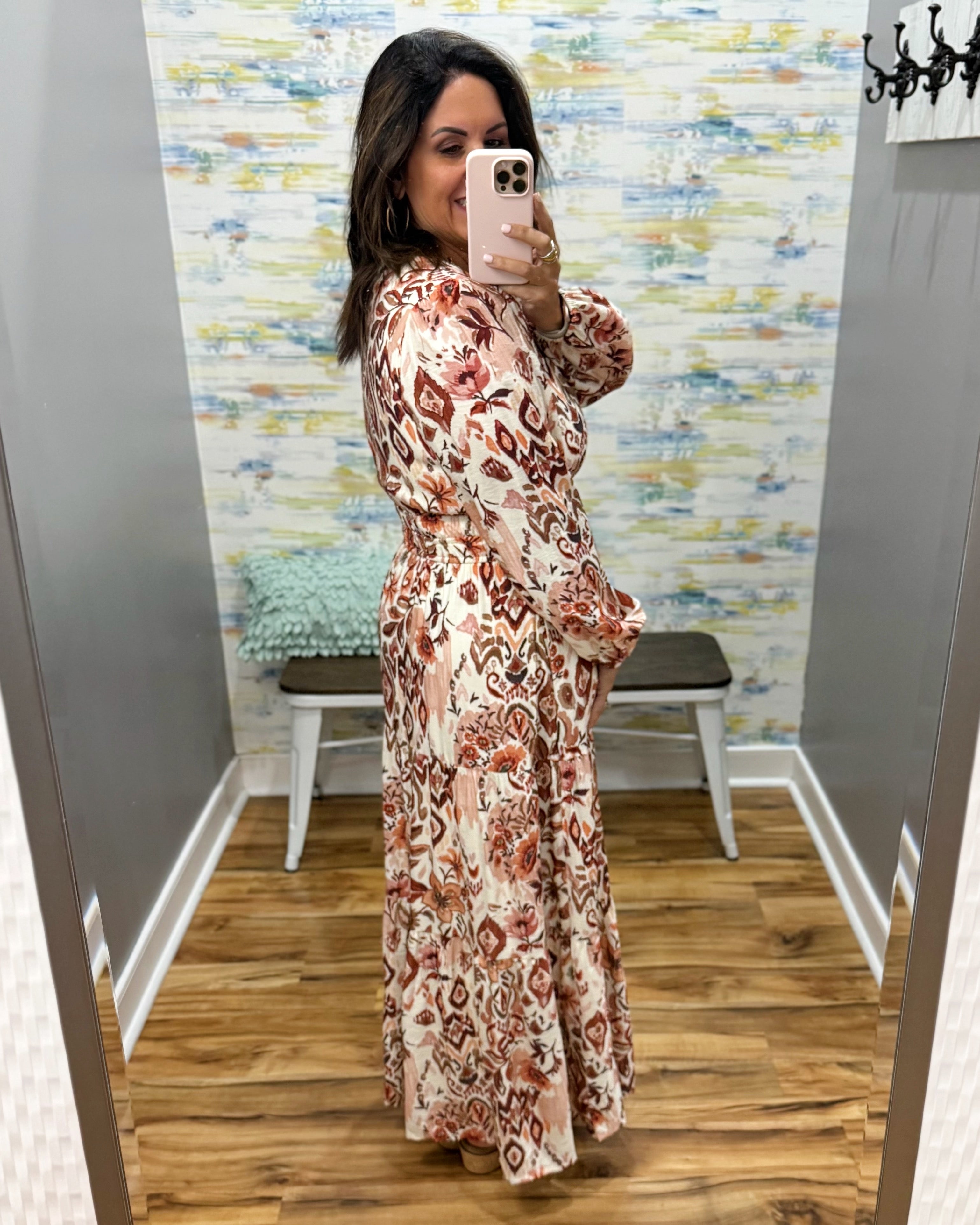A woman takes a mirror selfie in a fitting room wearing a long-sleeve maxi dress with a brown, rust, and beige abstract pattern on a cream background.