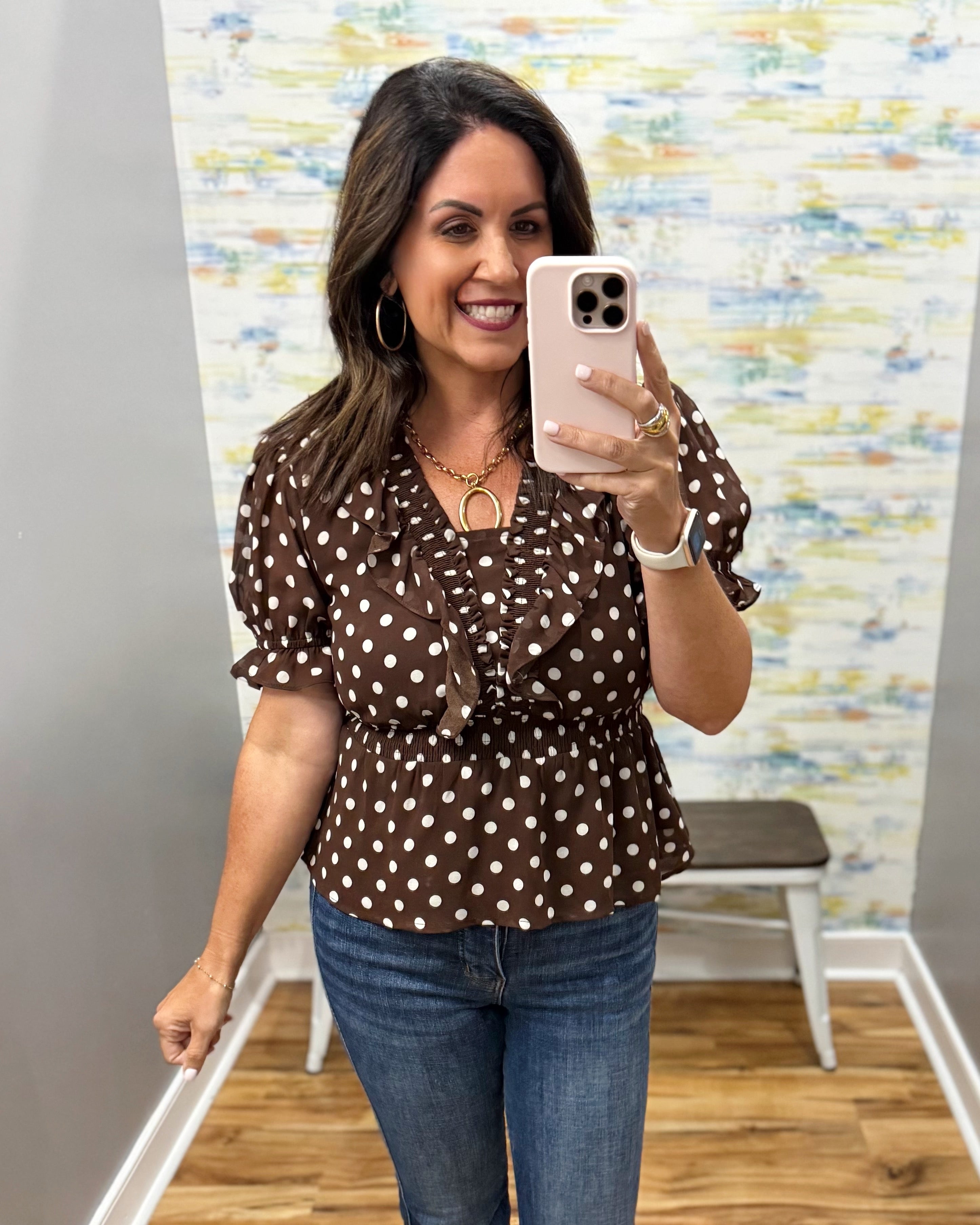 A woman taking a mirror selfie in a boutique wearing a brown short-sleeve blouse with white polka dots and blue jeans.