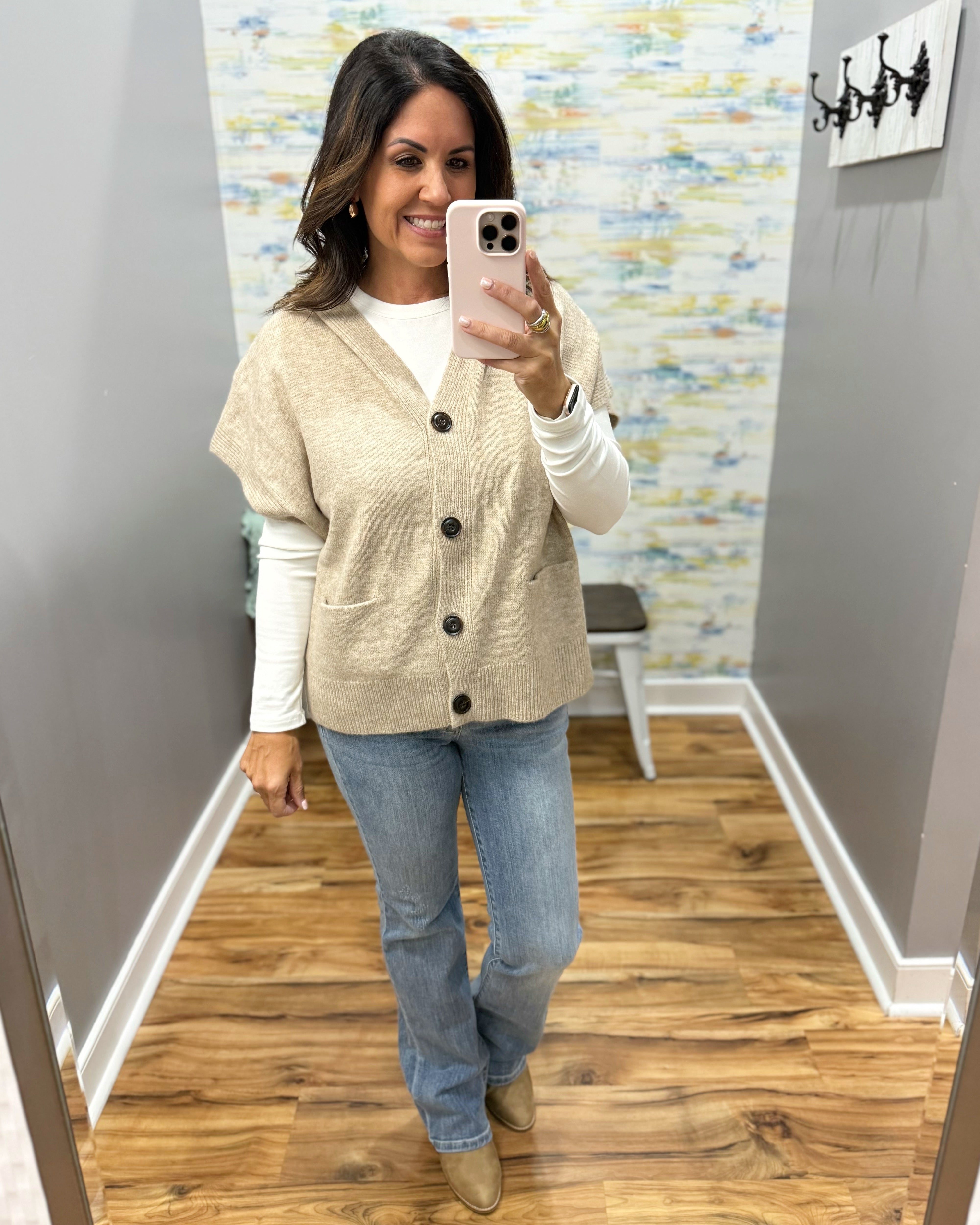 A woman takes a mirror selfie wearing a beige short-sleeve button-front cardigan over a white long-sleeve top, paired with light blue jeans and tan boots, in an indoor room with wood floors and gray walls.