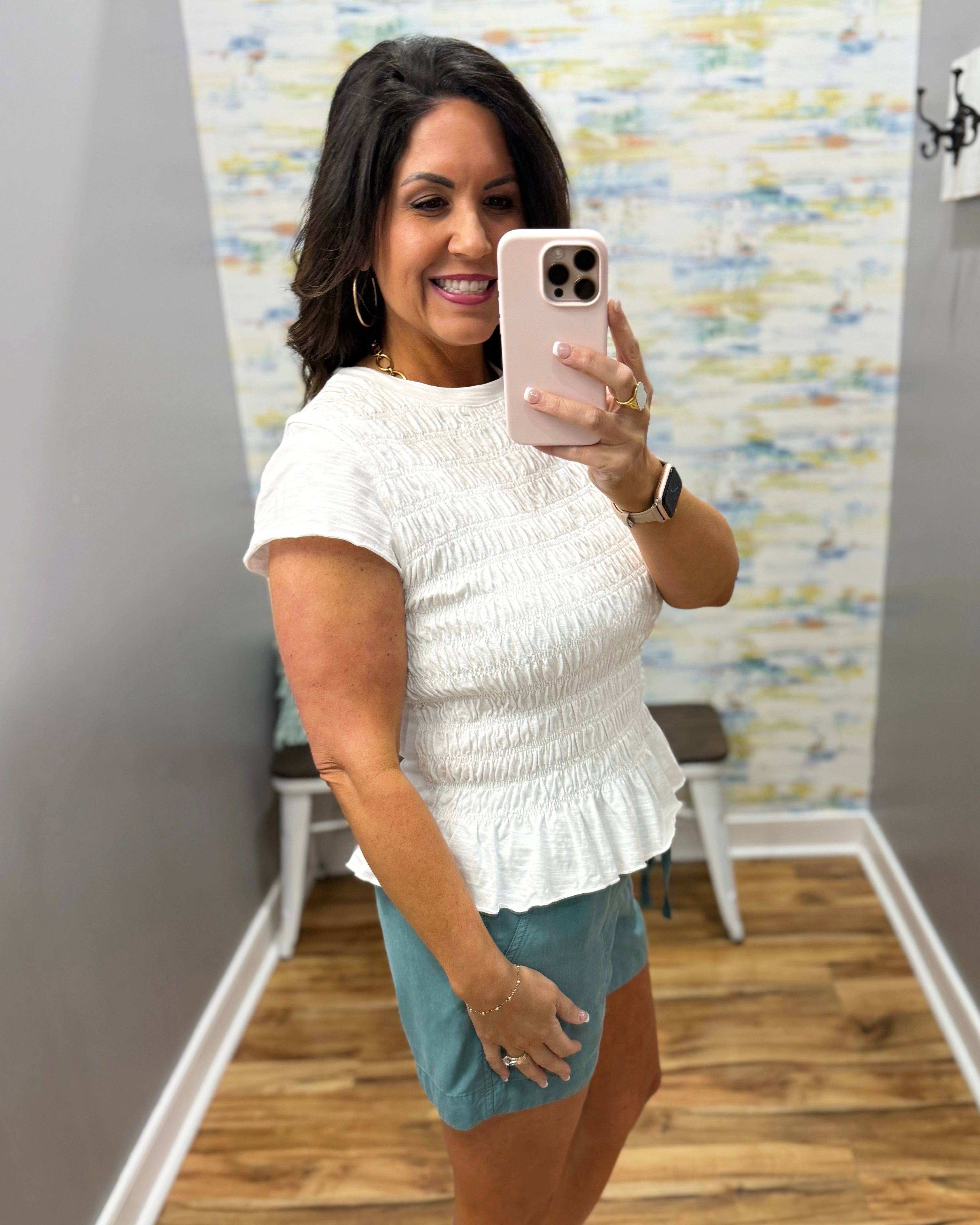 Woman taking a mirror selfie wearing a white textured top and blue shorts in a room with a colorful wall.