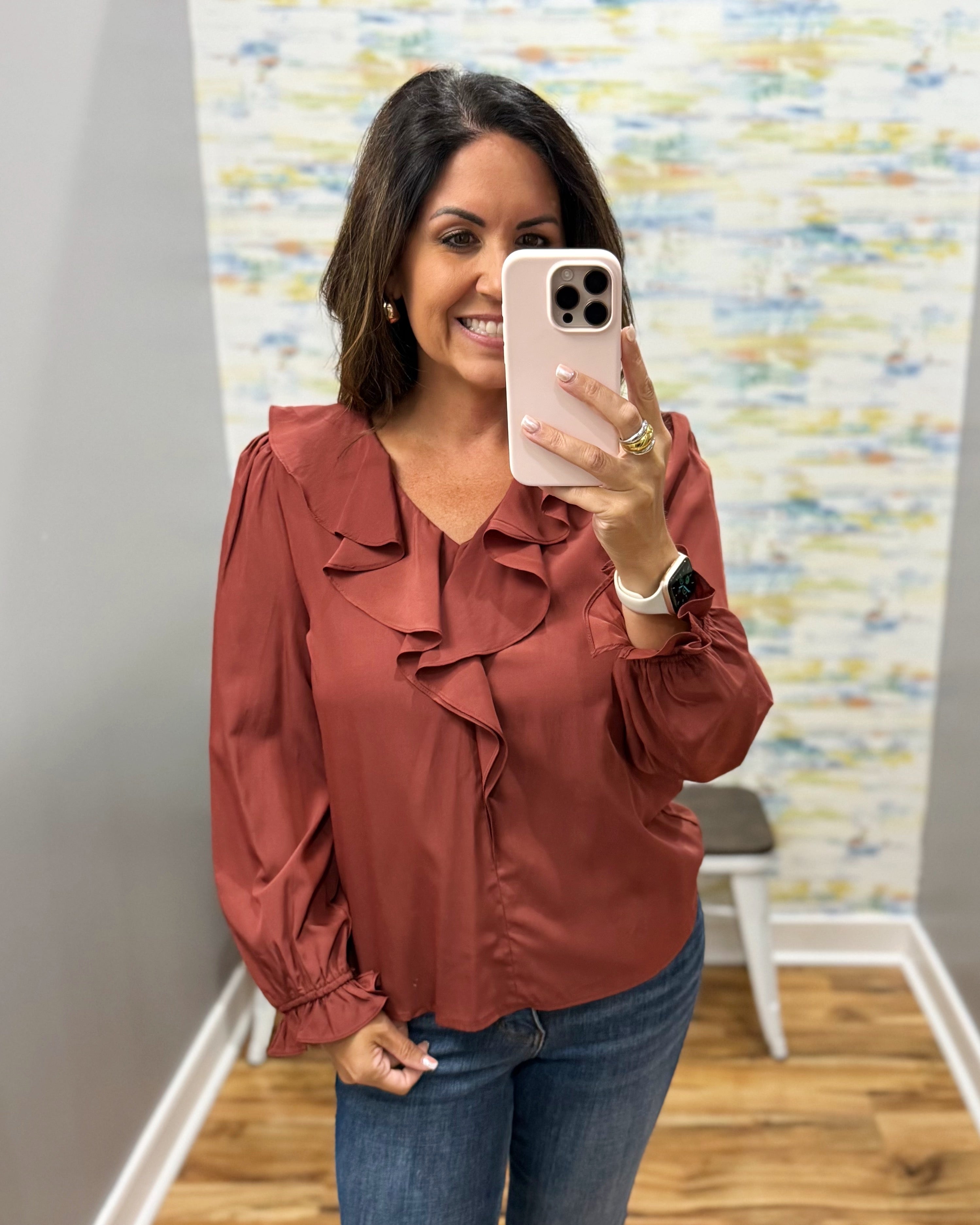 A woman takes a mirror selfie wearing a rust-colored long-sleeve blouse with ruffle details and blue jeans in an indoor setting.