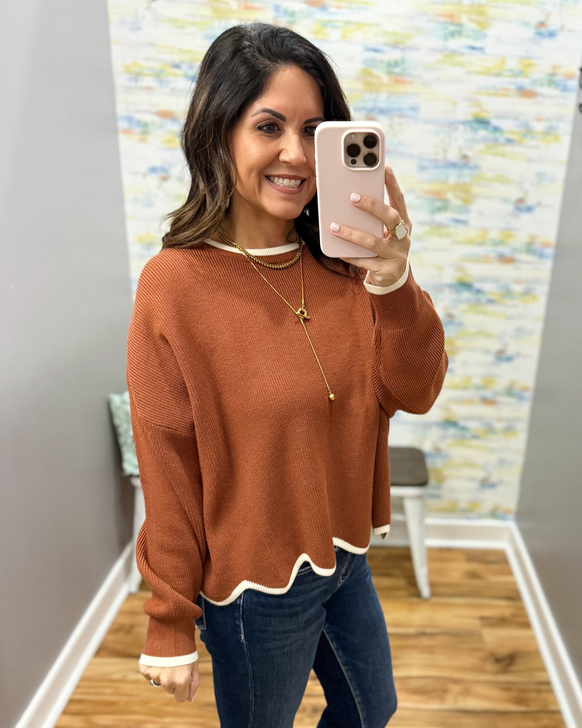 A woman takes a mirror selfie wearing a rust-orange knit sweater with white ribbed trim at the crew neck and scalloped hem, paired with dark blue jeans.